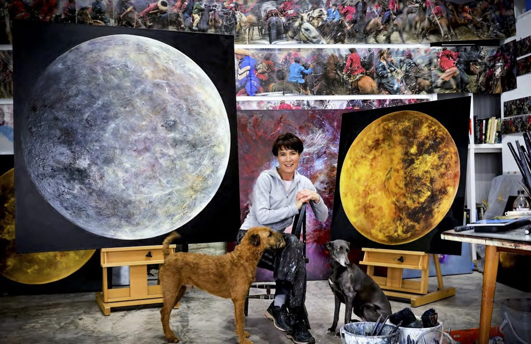 An artist in a studio displays two large paintings of moons, one grayish and the other yellowish, with two dogs sitting in front of her. There are additional artwork and supplies visible in the studio.
