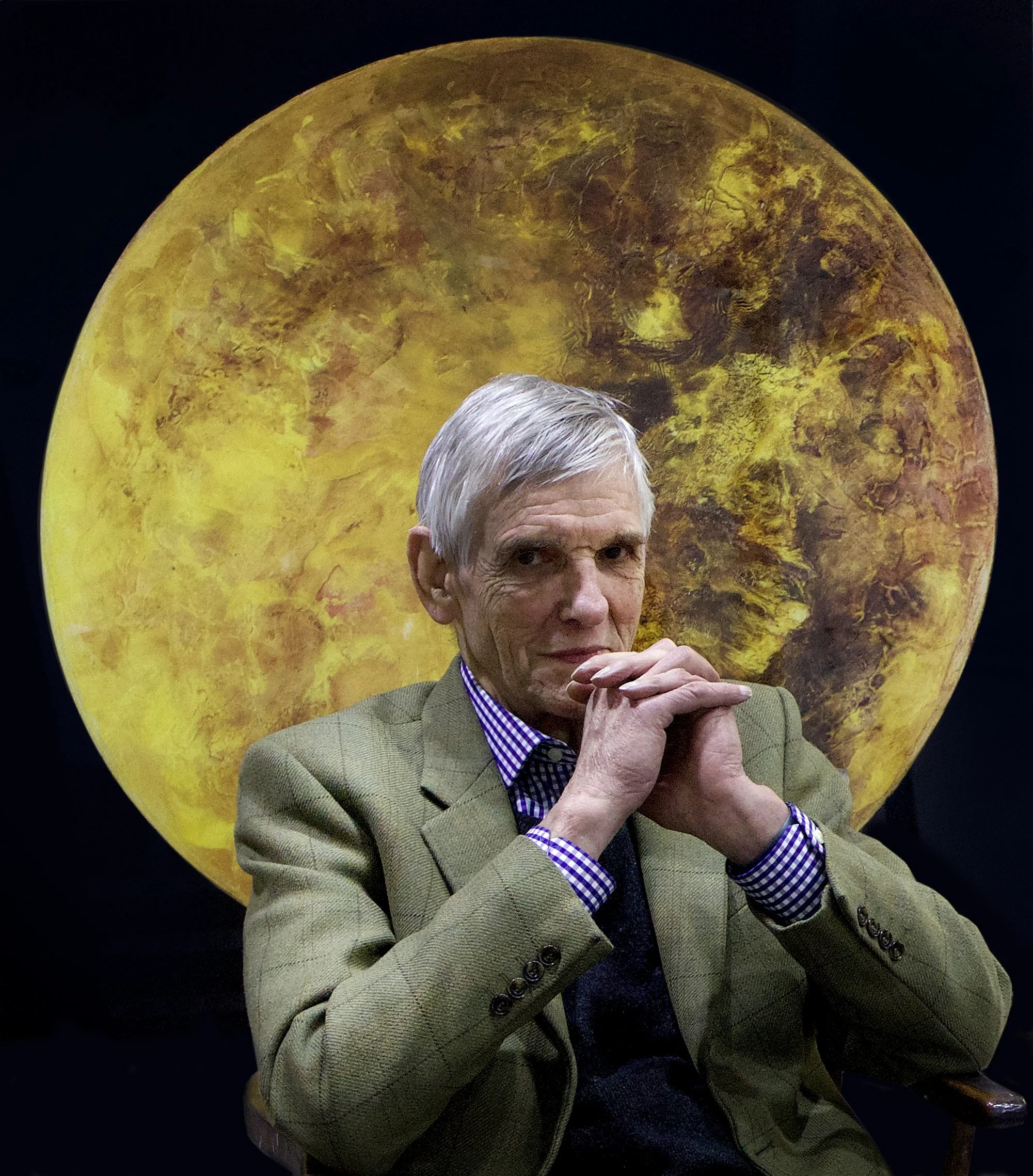 A man with gray hair, wearing a brown checked blazer and purple checkered shirt, is sitting with his hands clasped in front of his face. Behind him is a large artwork resembling a yellow-orange planet.