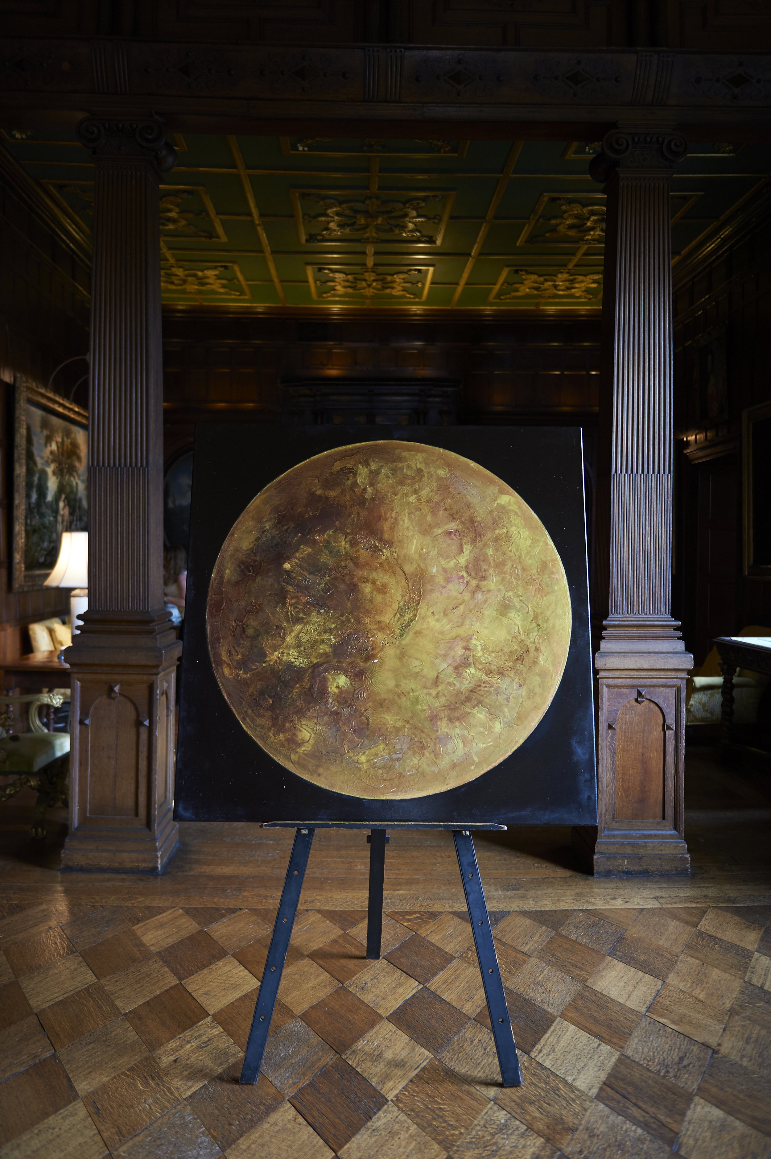 A painting of the moon on an easel in a room with wood paneling and ornate ceiling, with columns framing the scene.