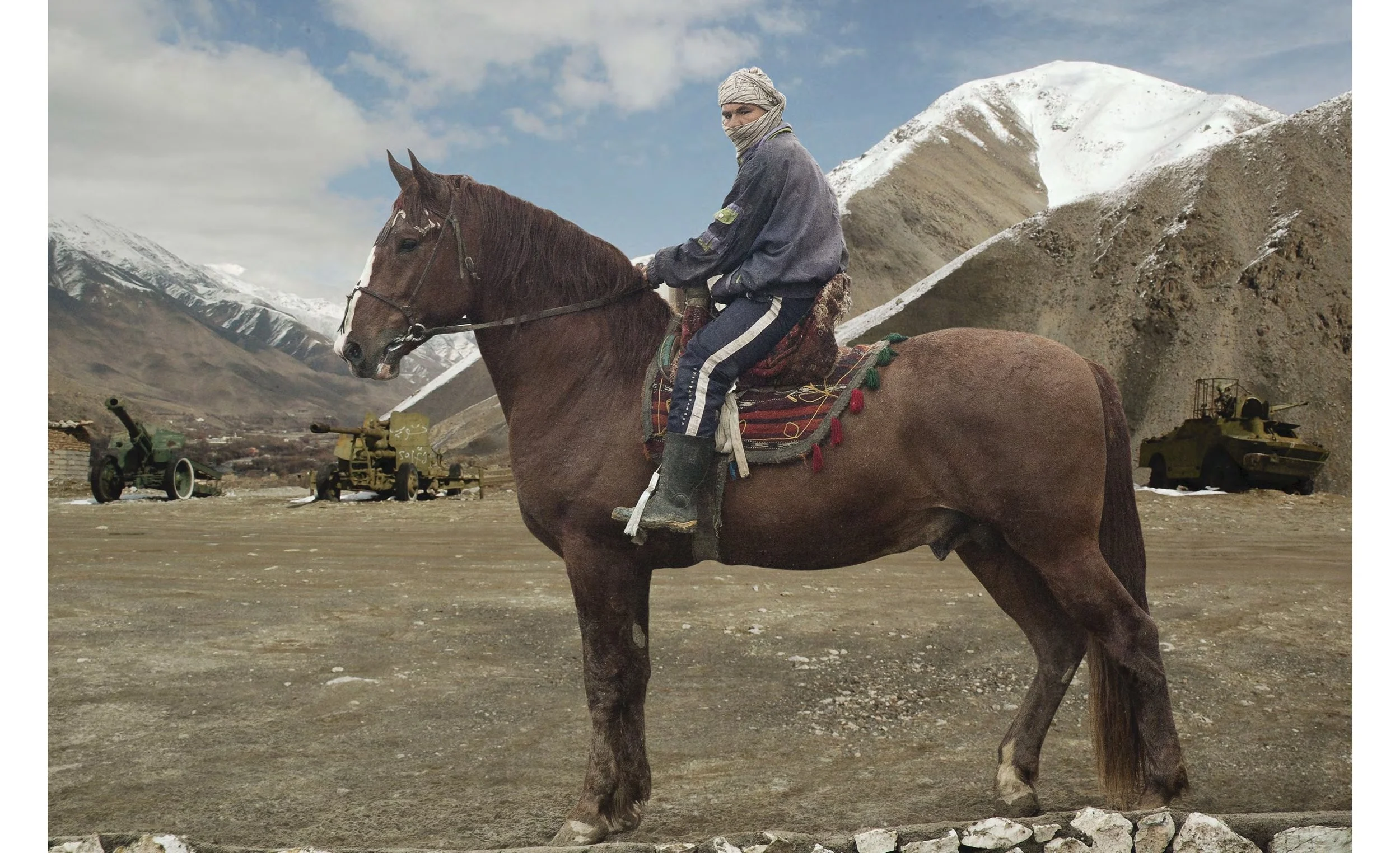 A person riding a brown horse in a mountainous landscape with snow-capped peaks and military tanks in the background.
