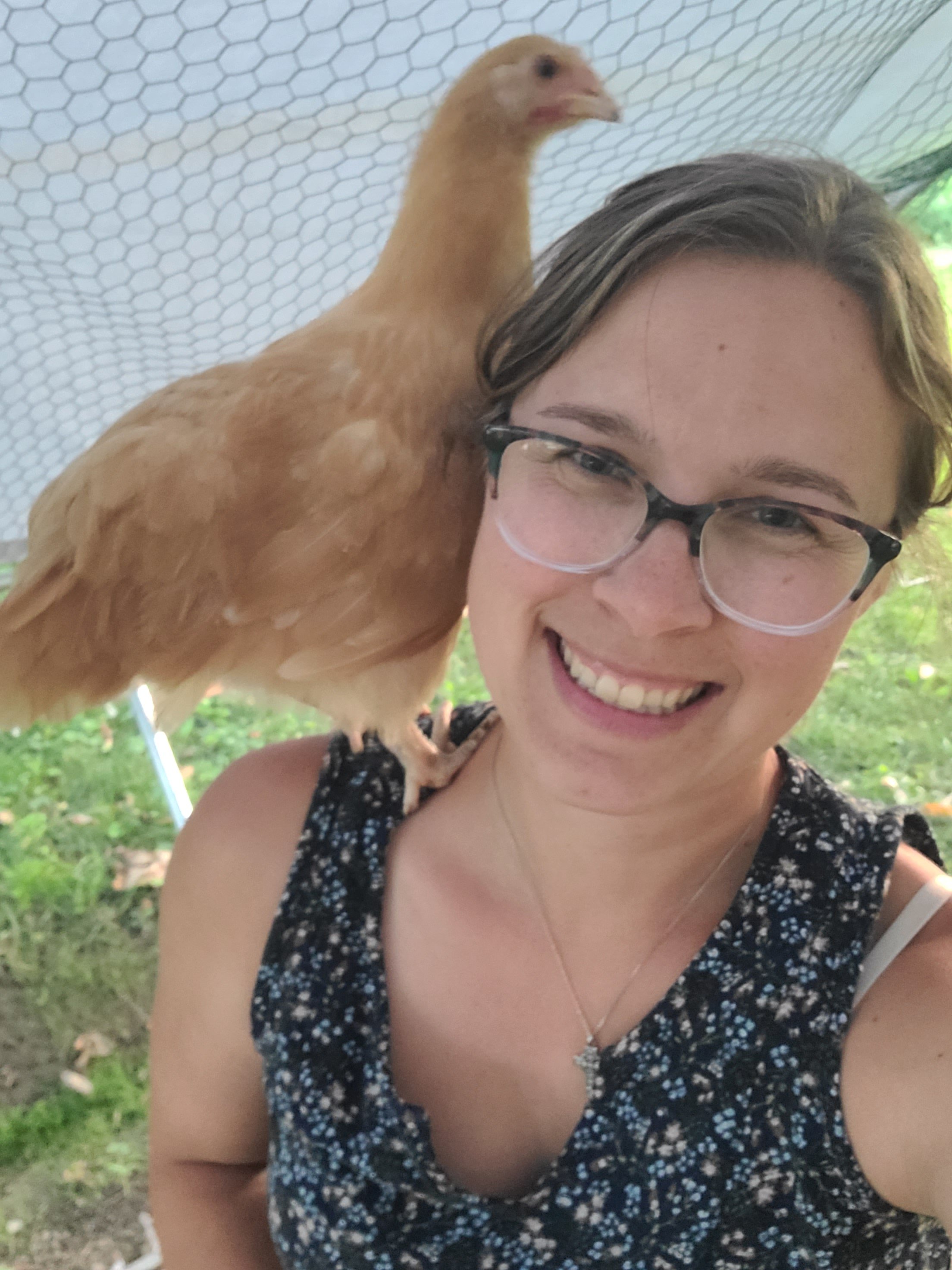 A woman with glasses smiling, standing outdoors with a chicken perched on her shoulder.