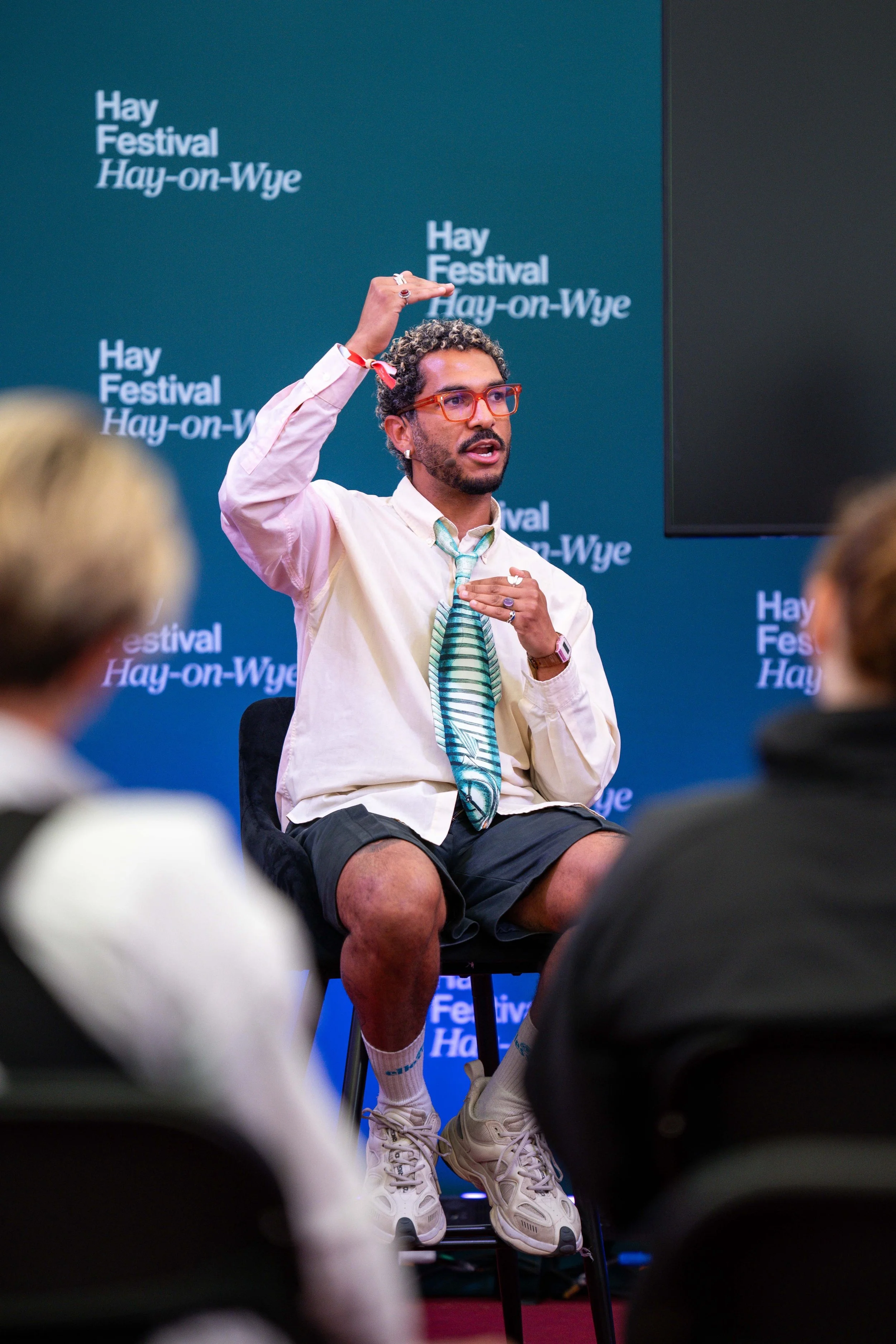 A man sitting on a chair in front of a blue backdrop with repeated white text that reads "Hay Festival Hay-on-Wye." He is speaking, gesturing with his right hand, and has one hand on his chest. He is wearing a light-colored, long-sleeved shirt, a turquoise and white striped tie, dark shorts, white socks, and white sneakers. He has curly hair, glasses, and a beard.