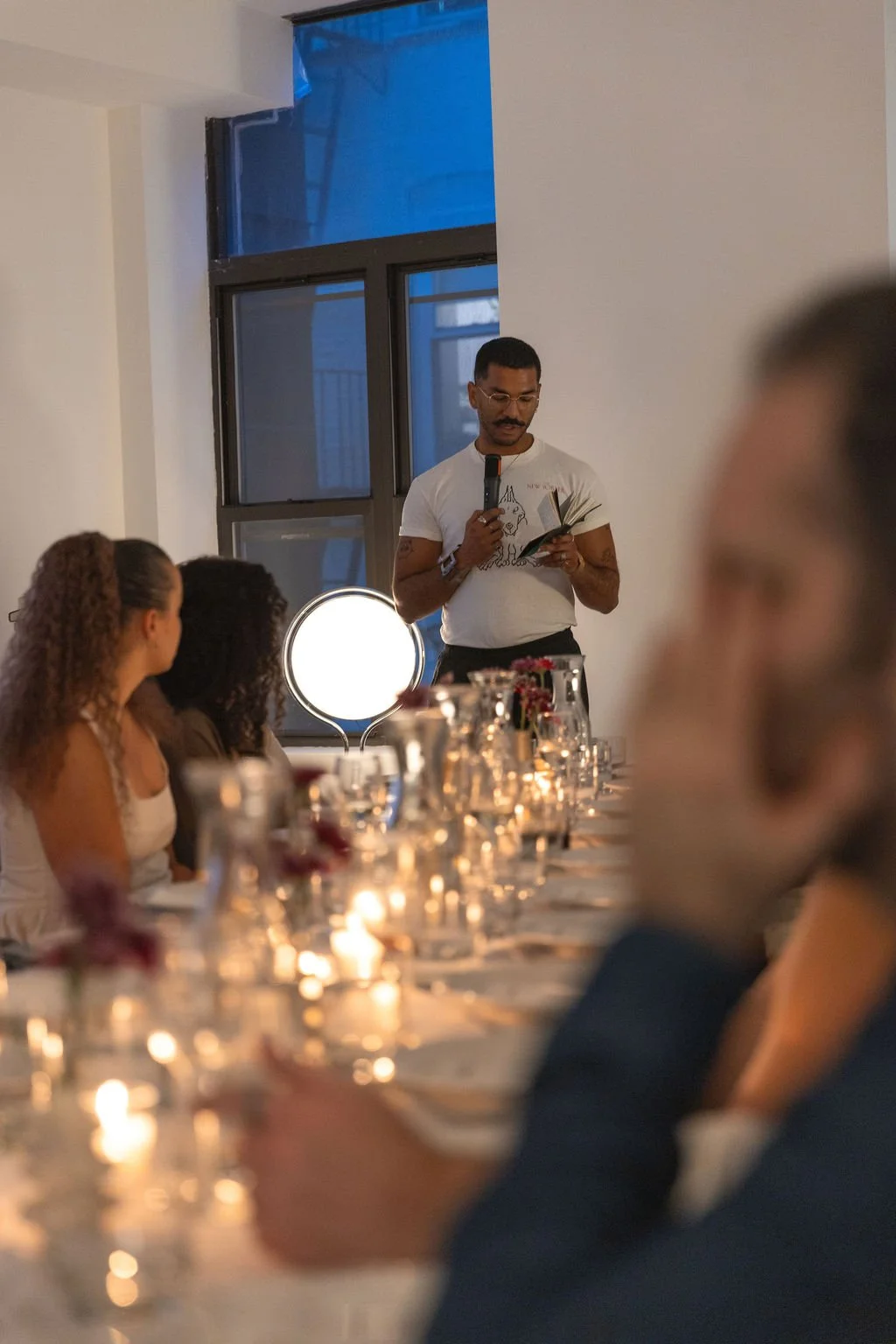 A man with a mustache and glasses is standing at a table, reading from a book into a microphone, in a warmly lit indoor setting. Several people are seated around the table, listening attentively. The table is decorated with candles and flowers, creat