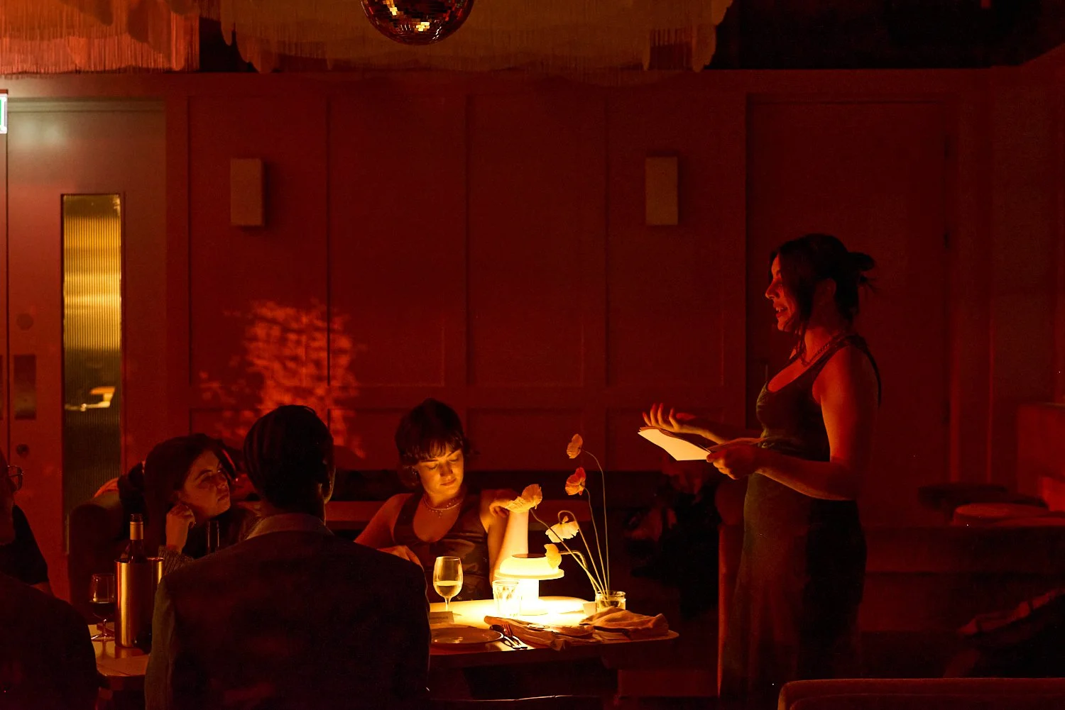 A woman in a black dress standing and speaking with a paper in her hand, while five people sit around a table in a dimly lit restaurant or bar with red lighting, drinks, and a floral centerpiece.