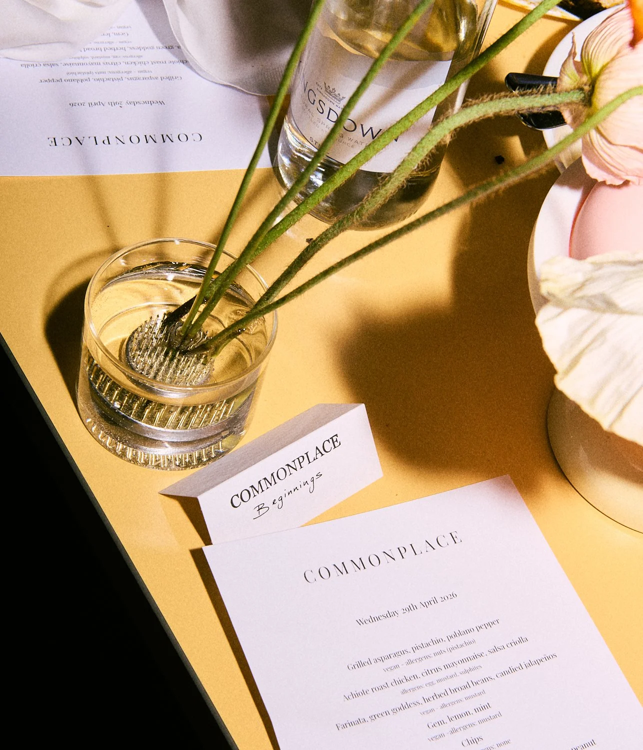 A glass with water and green plant stems, a printed menu titled "COMMONPLACE" with the subtitle "Beginnings", a paper with the same title and date, a bottle of water, and a flower arrangement on a yellow table.