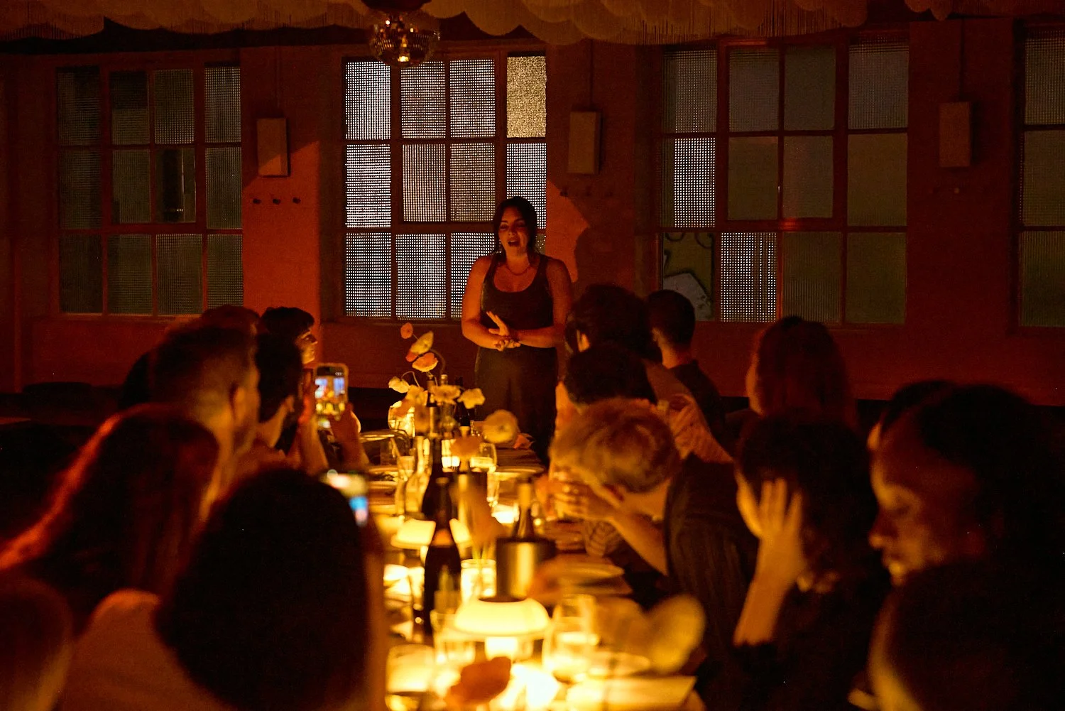 Woman standing and speaking in front of seated audience during a dinner event in a dimly lit room decorated with flowers and warm lighting.