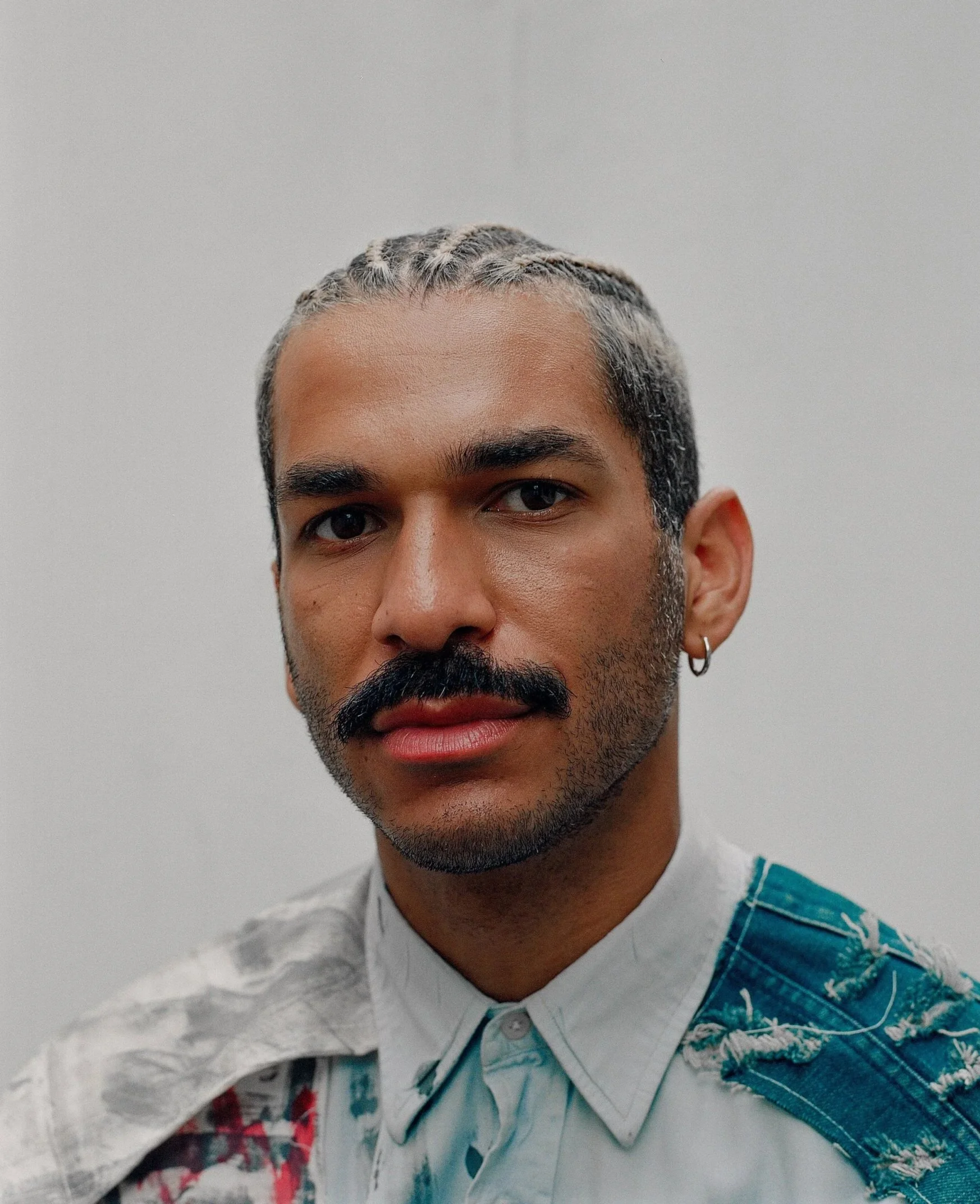 Partial view of a young man's face with dark hair, beard, and earring, wearing a white shirt and a blue plaid shirt over it, standing against a plain light-colored background.