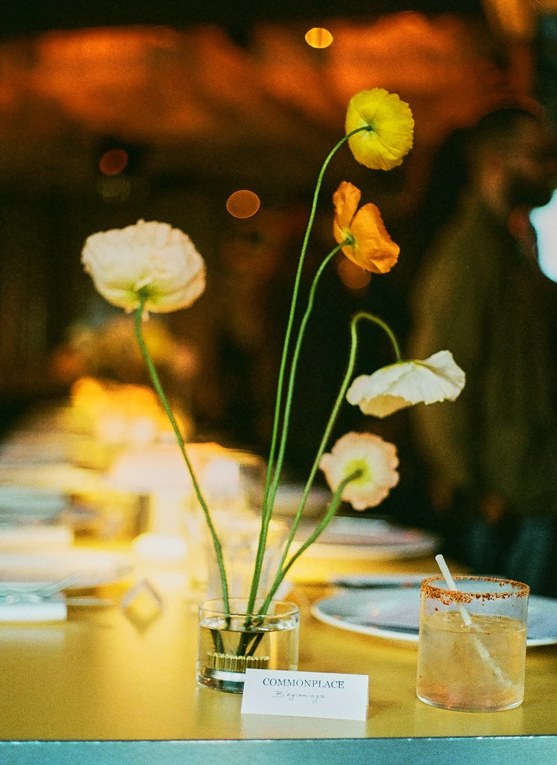 Vase with tall, delicate flowers in cream, white, and orange, set on a table with out-of-focus background and a small card labeled 'COMMONPLACE Beginnings'.