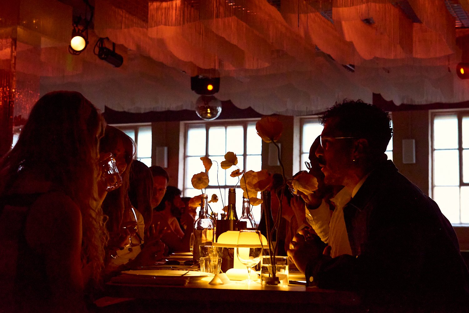 People dining at a restaurant table with dim lighting, decorative flowers, and a glowing lamp.