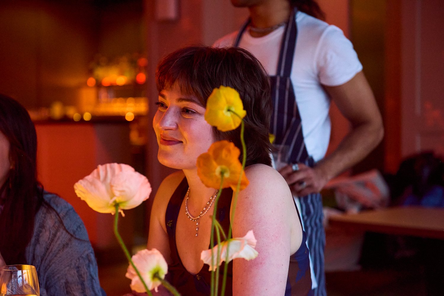 A woman with short brown hair and freckles smiling at a table with colorful flowers in the foreground, in a dimly lit dining setting, with a person in a white shirt and striped apron in the background.