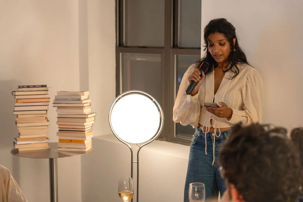 A woman holding a microphone and reading from her phone at a book launch or event, with stacks of books on a table nearby and a bright light illuminating the scene.