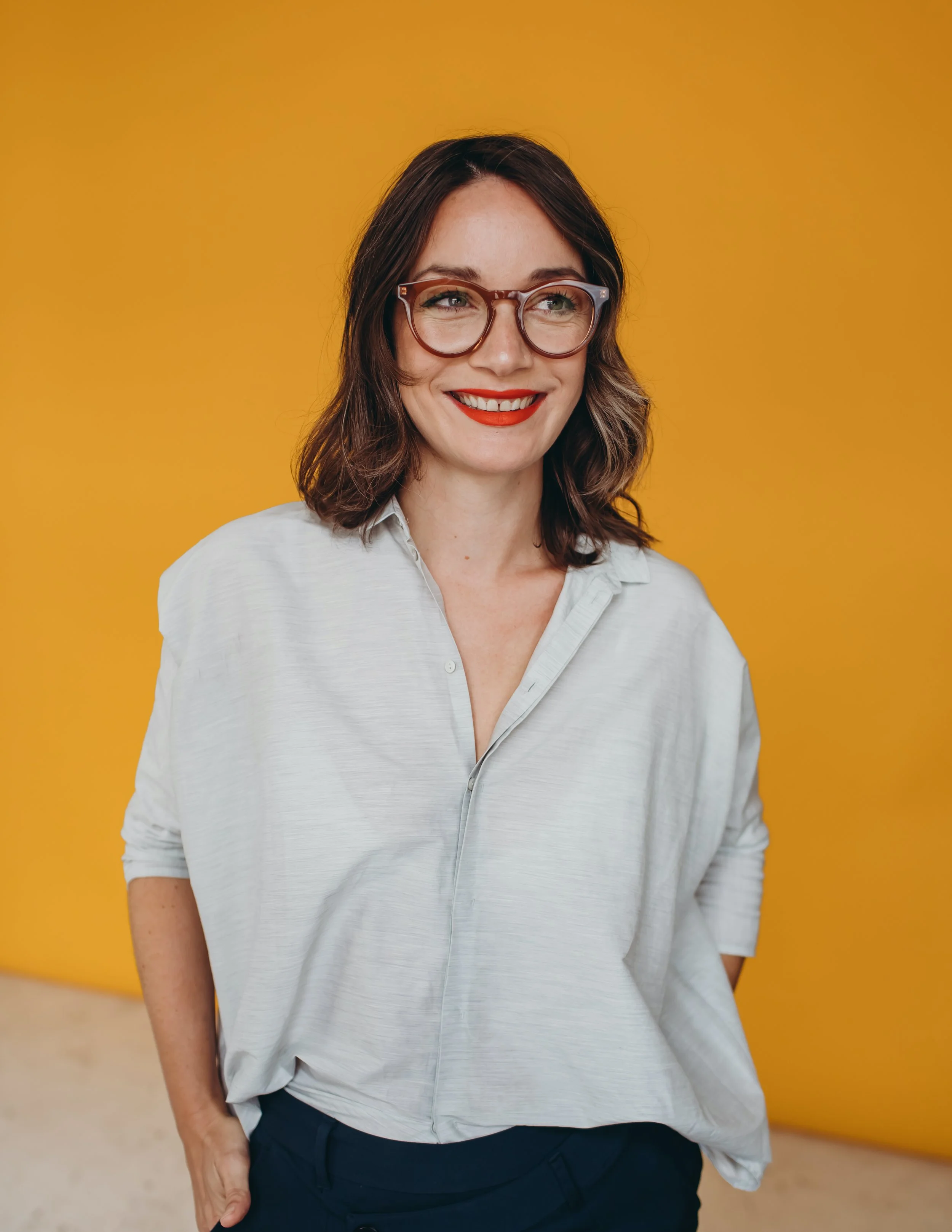 A woman with wavy brown hair and glasses, wearing a white shirt, standing against a yellow background and smiling.