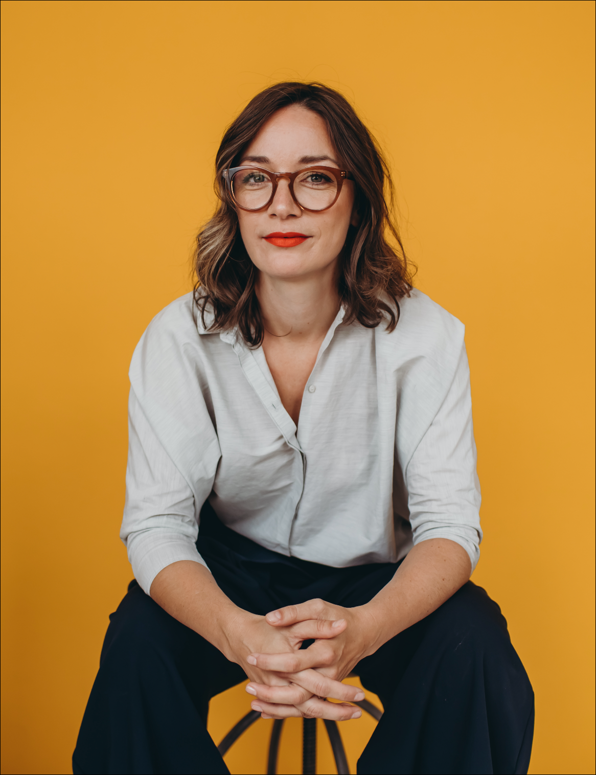 A woman with shoulder-length brown hair, wearing glasses, a white button-up shirt, and dark pants, sitting against a yellow background.