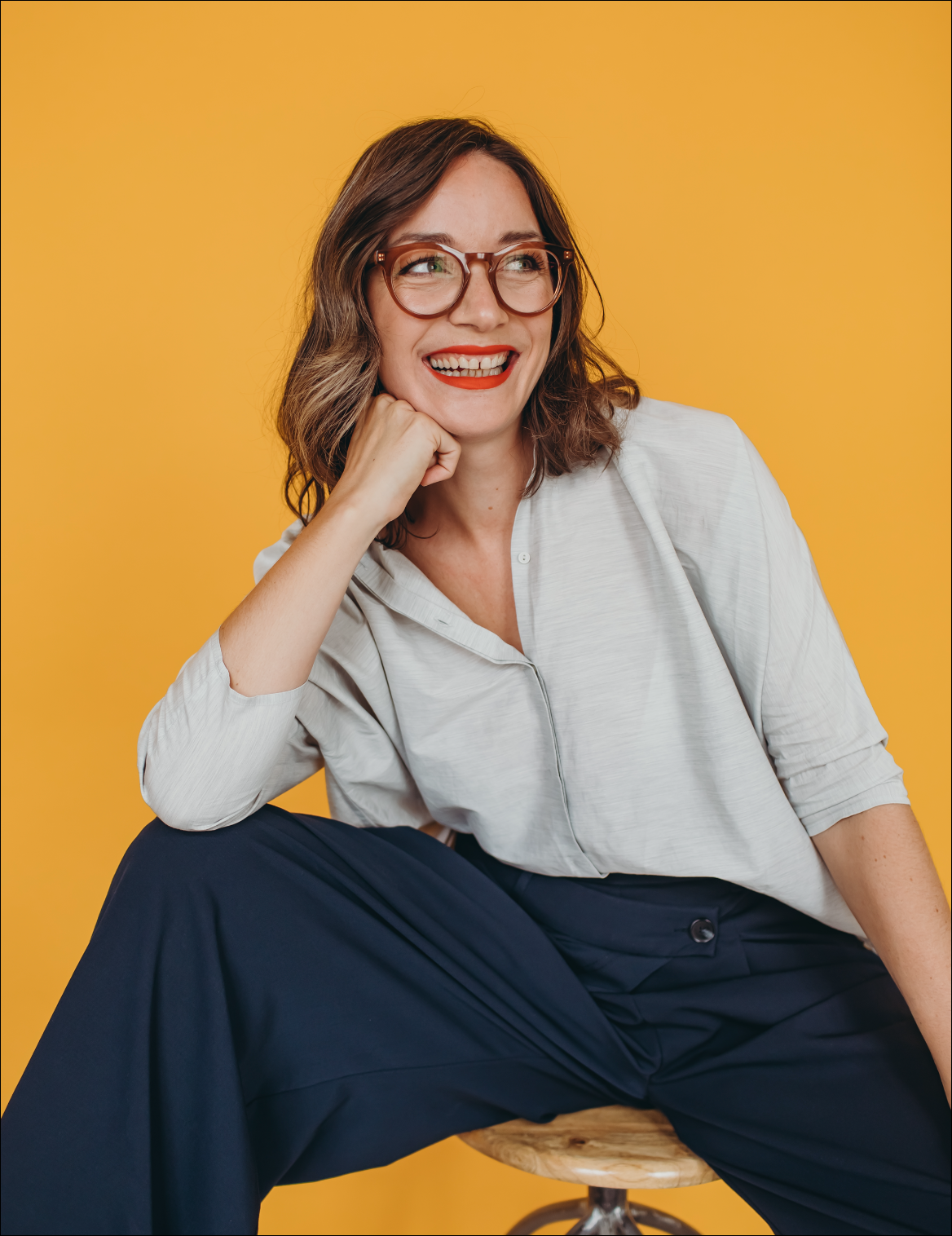 A woman with brown hair, glasses, and red lipstick sitting on a stool with a yellow background. She is smiling and wearing a light-colored blouse and dark pants.