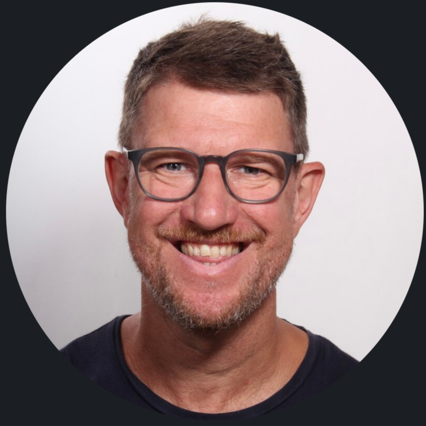 Headshot of a smiling man with glasses, short brown hair, and a beard, wearing a dark shirt against a white background.