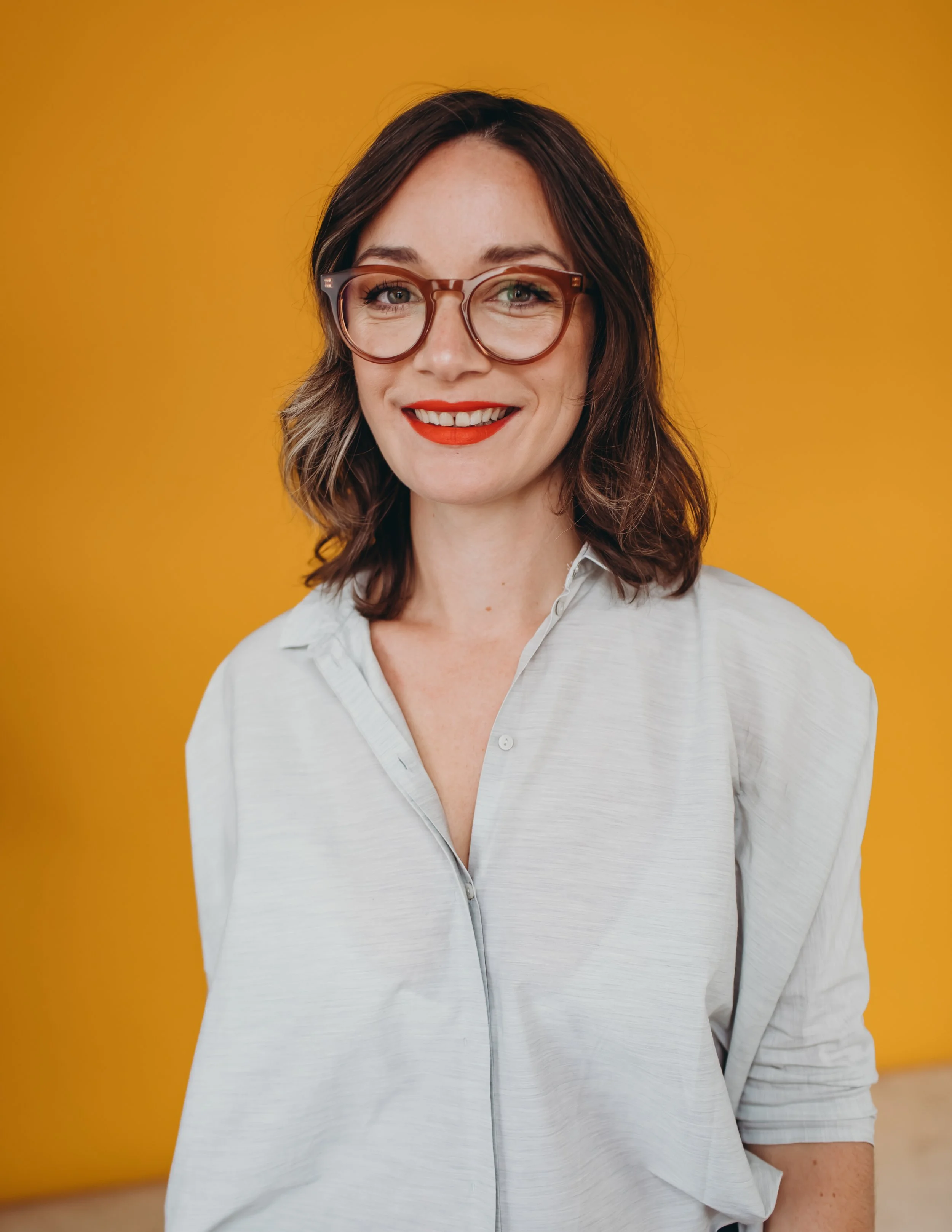 Portrait of a woman with shoulder-length brown hair, wearing large round glasses, red lipstick, and a light-colored button-up shirt, standing against a solid yellow background.