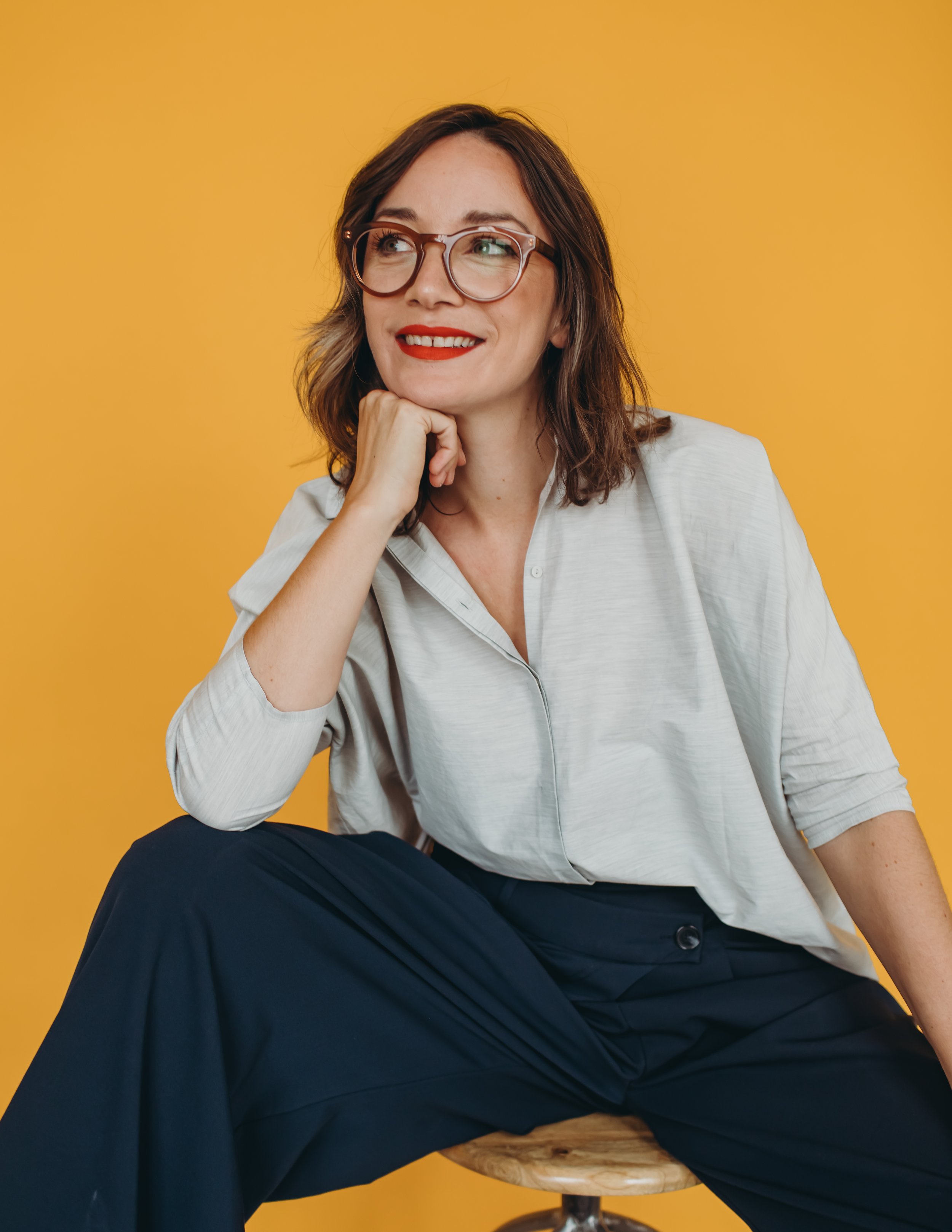 A woman with shoulder-length brown hair, wearing glasses, a white shirt, and dark pants, sitting on a wooden stool against a plain yellow background, smiling and looking to the side with her hand resting on her chin.
