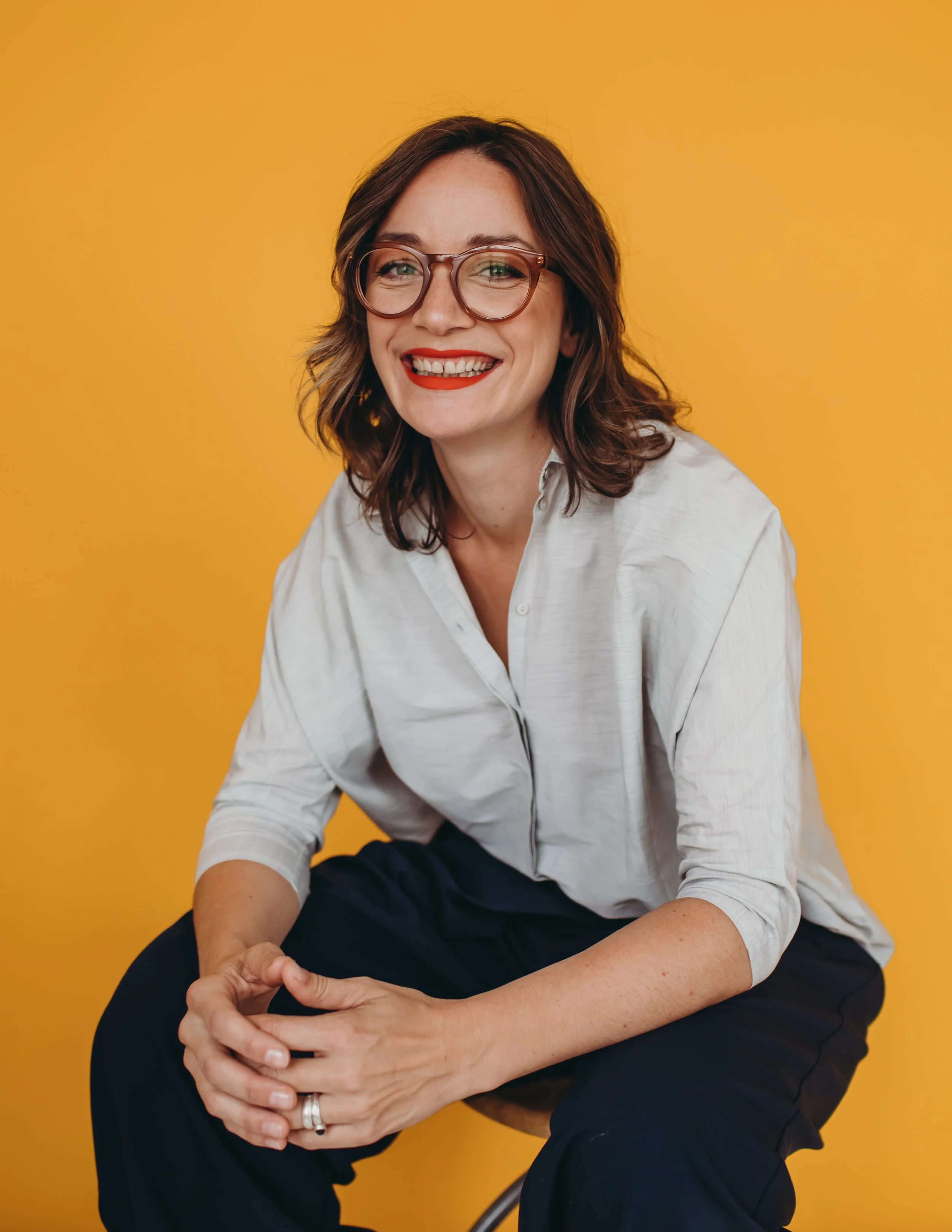 A woman with shoulder-length brown hair, glasses, and red lipstick, smiling while sitting against a yellow background.