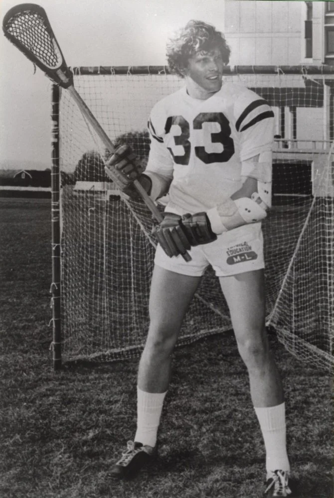A young person with curly hair wearing a sports jersey numbered 33, shorts, and gloves, standing on a field with a lacrosse stick in hand and a goal net behind them.