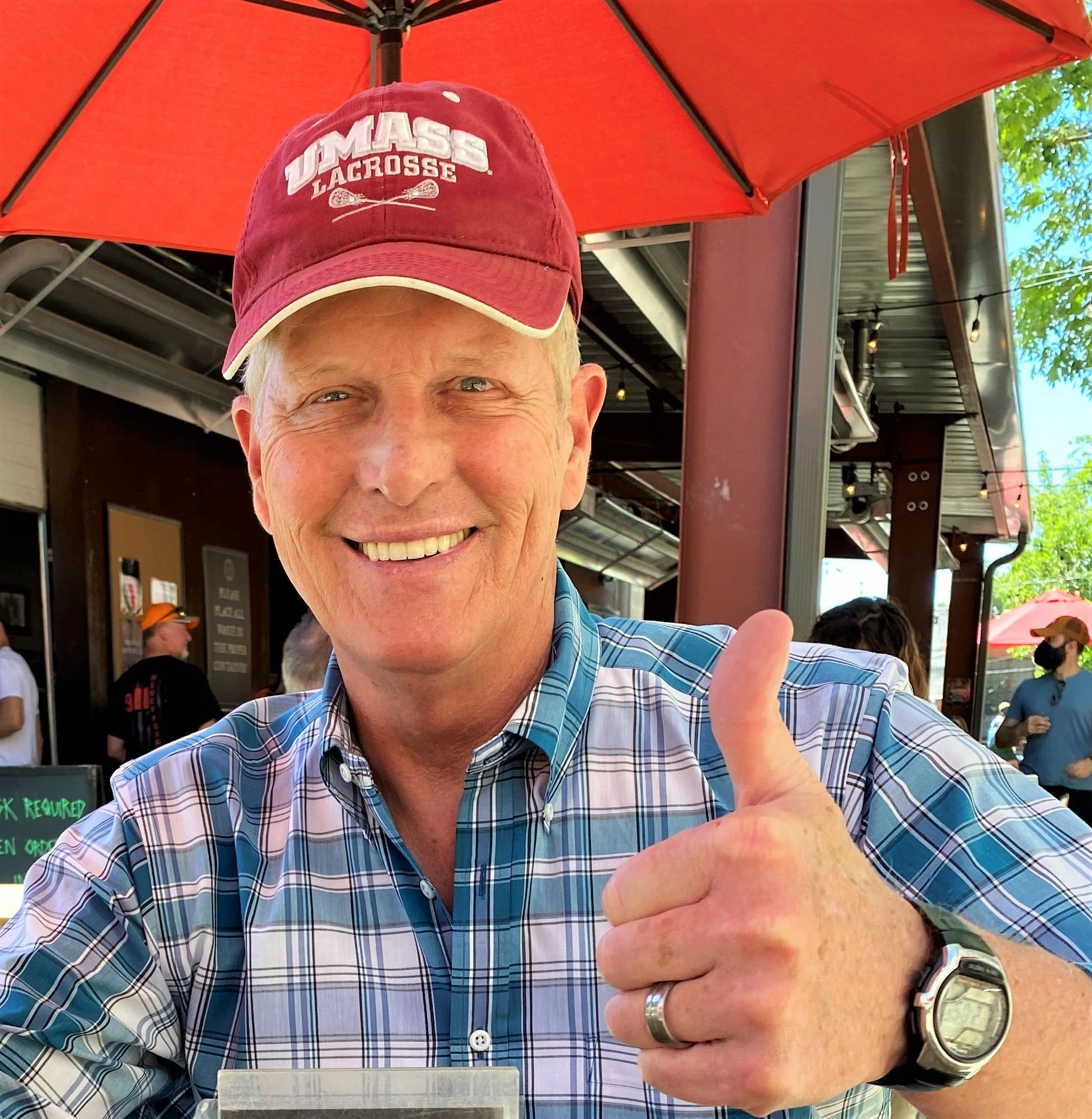 A smiling man wearing a red cap with white lettering that says "UMASS Lacrosse" and a blue plaid shirt. He is giving a thumbs-up in an outdoor setting with a red umbrella and other people in the background.
