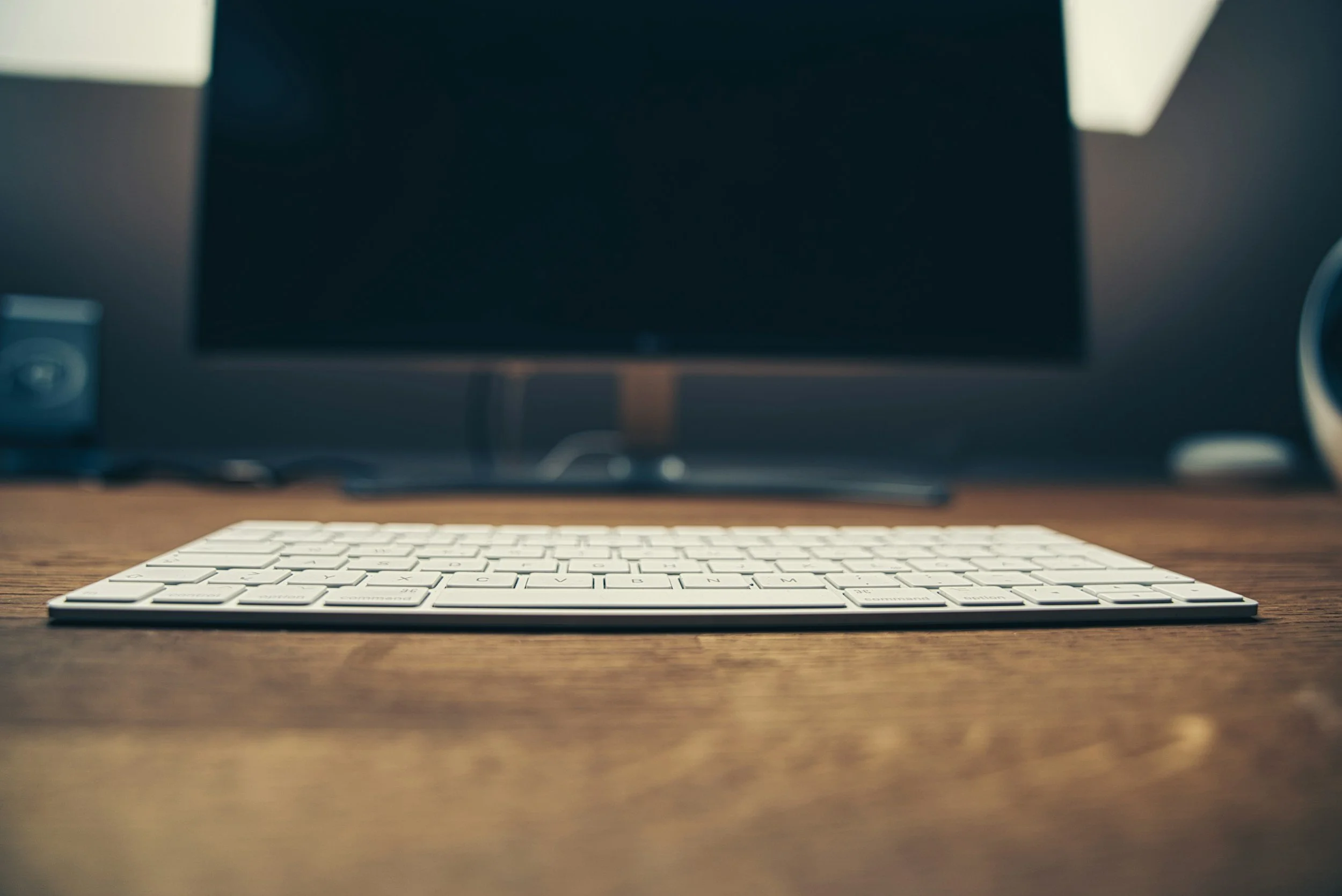 Close-up of a white computer keyboard on a wooden desk, with a large monitor and other electronic devices in the background.