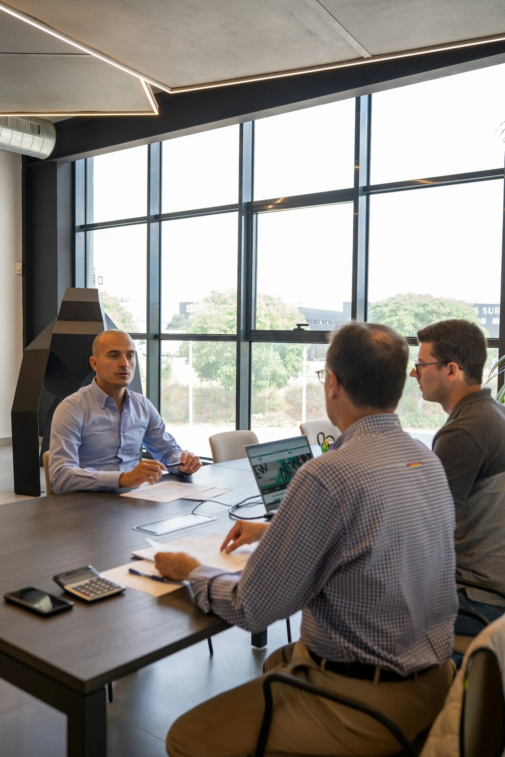 A business meeting taking place in a conference room with large windows, with four men seated around a table. One man, dressed in a light blue shirt, is speaking or presenting, while the others listen, some working on laptops and taking notes.