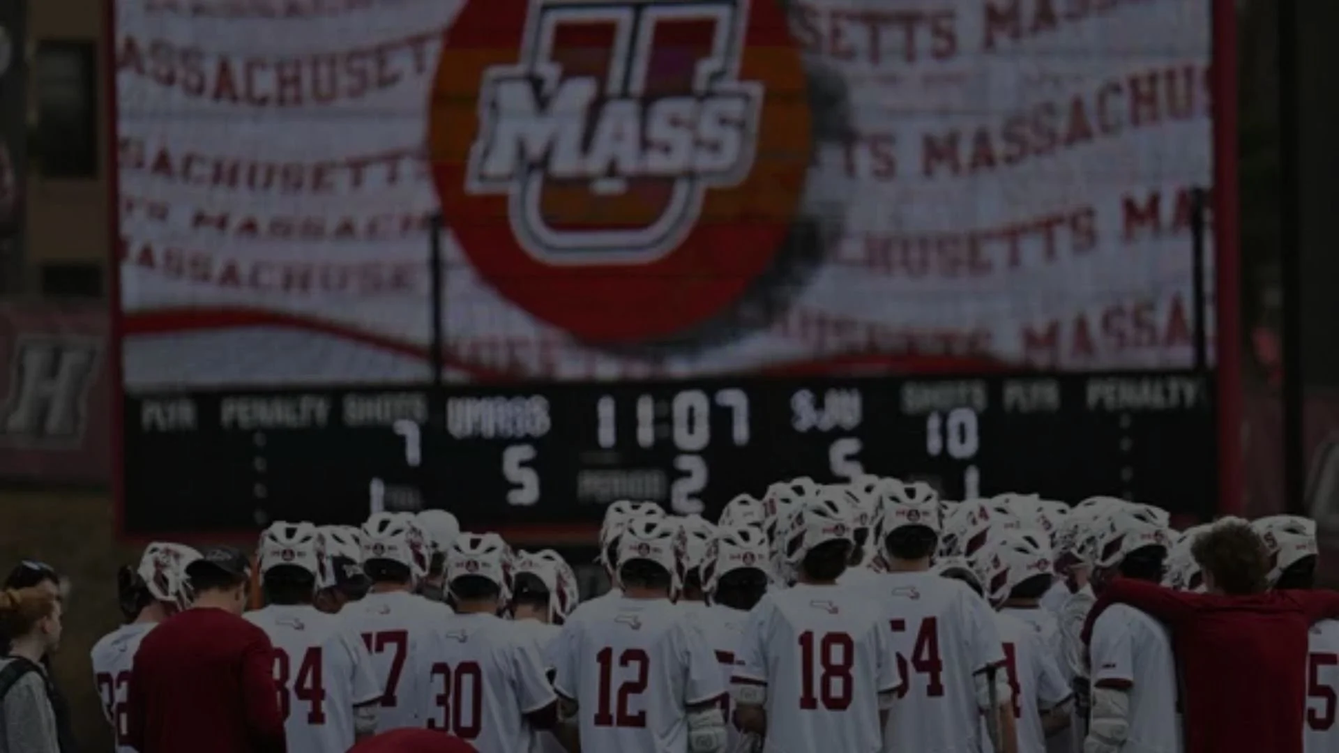 A group of lacrosse players in white jerseys and helmets standing in front of a scoreboard and a large banner.