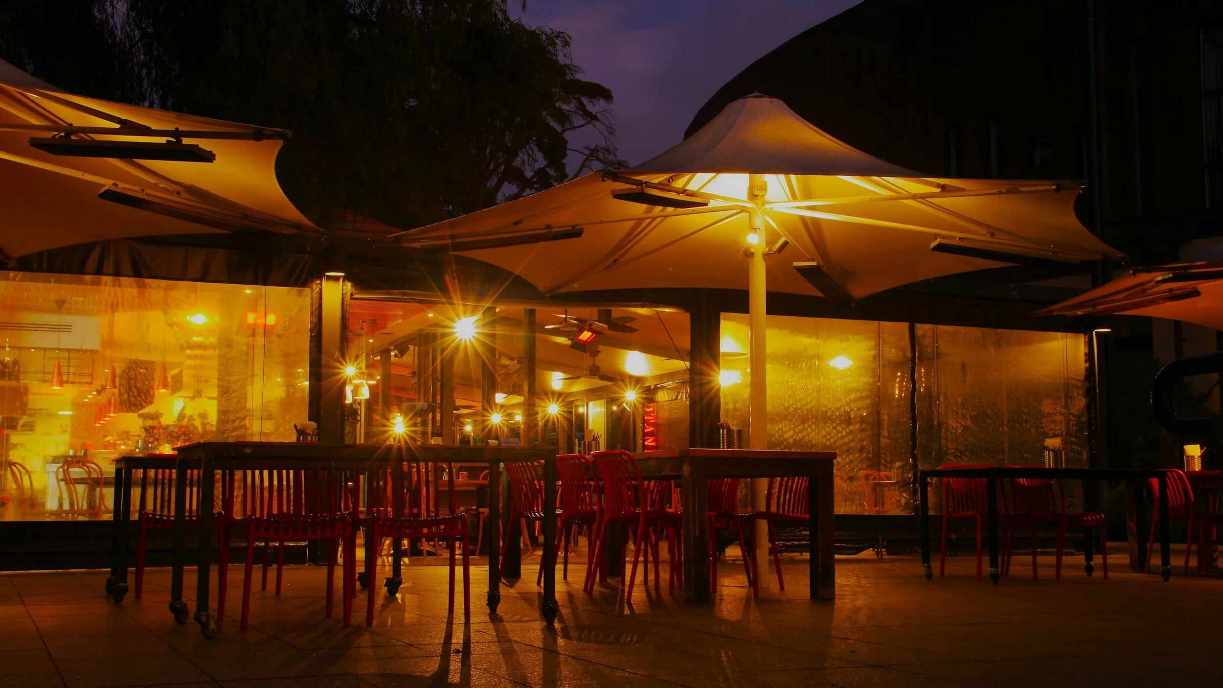 Nighttime view of an outdoor seating area with red chairs, illuminated by warm yellow lights and umbrellas.