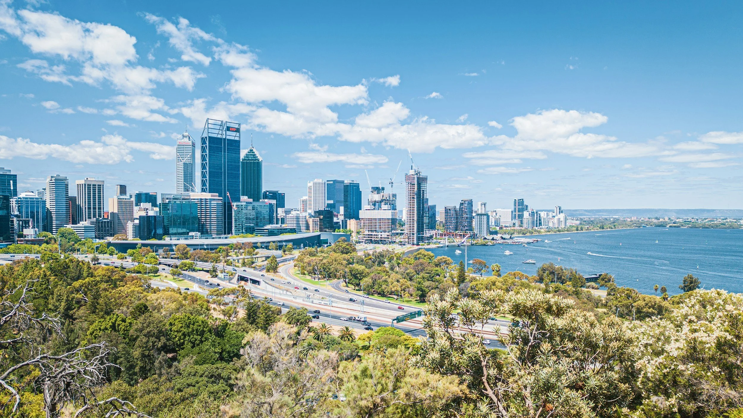 A city skyline with tall skyscrapers next to a large body of water, seen from a green park area with trees and a road in the foreground, under a blue sky with scattered clouds.