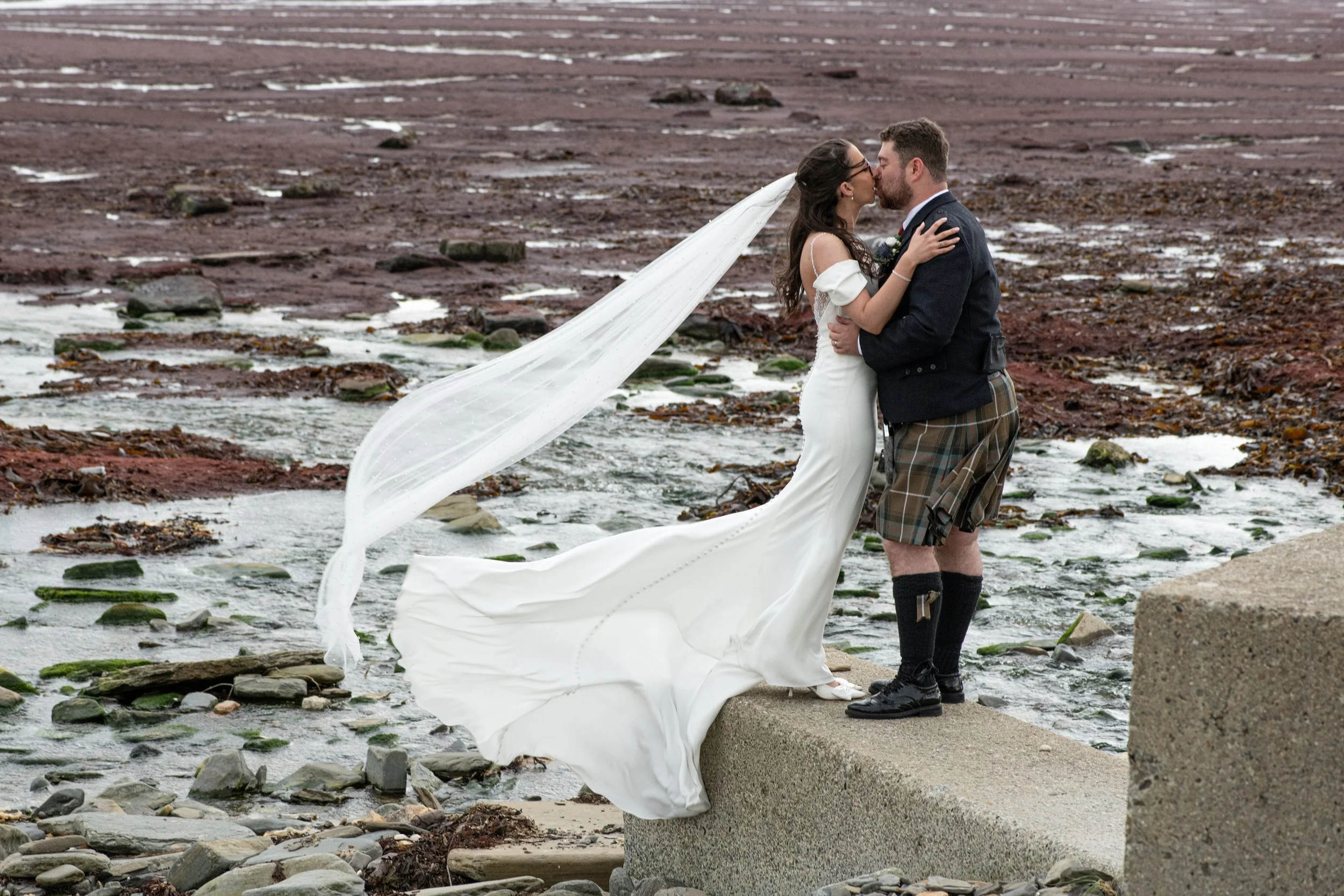A bride and groom standing on a rocky beach, sharing a kiss. The bride is wearing a white wedding dress with a long veil, and the groom is dressed in a dark jacket and traditional Scottish kilt with knee-high socks.