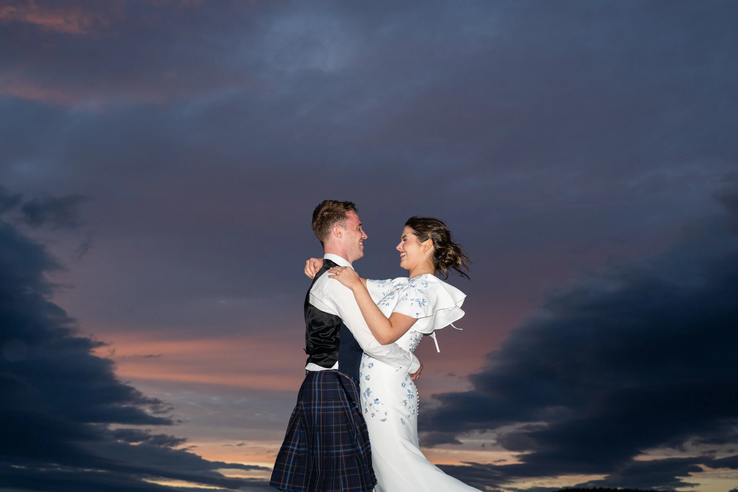 A couple dressed in wedding attire dancing outdoors at sunset, with a dramatic sky in the background.