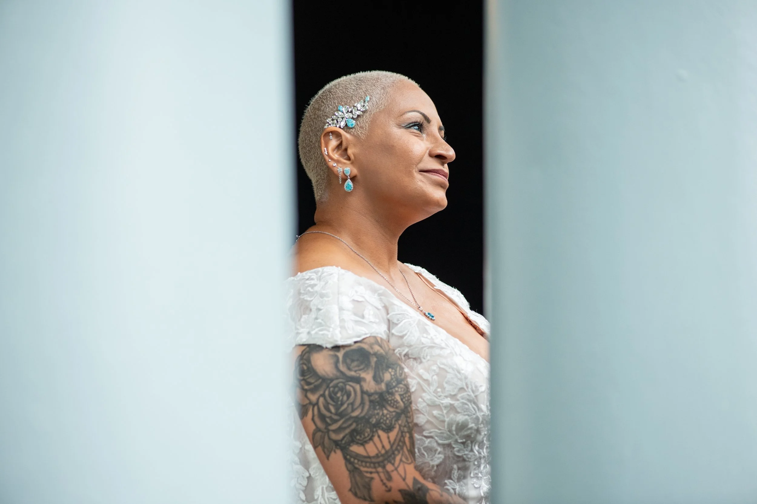 A woman with a short blonde haircut and tattoos on her arm, wearing turquoise jewelry and a white lace dress, looks to the side through a partially open door