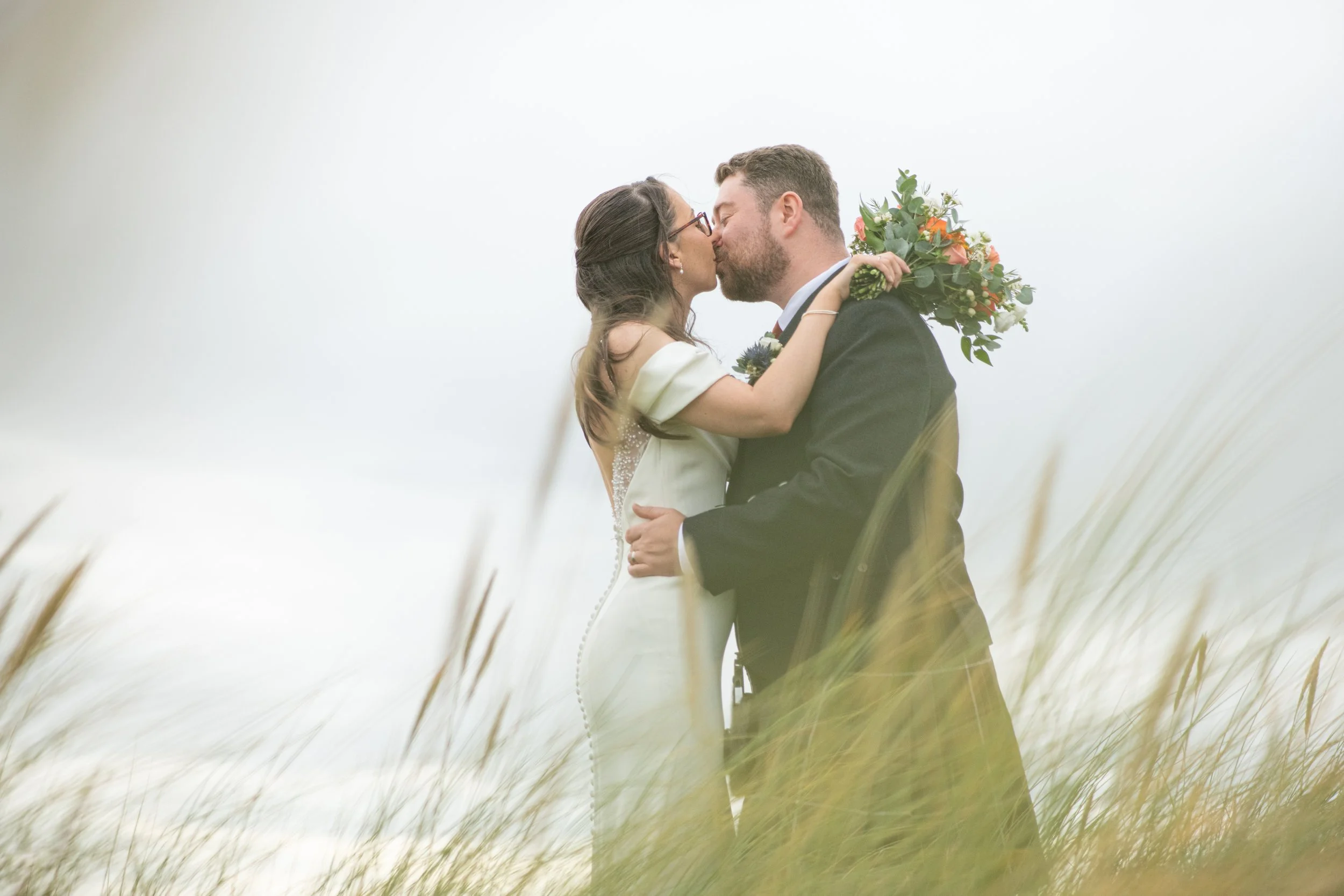 A bride and groom kiss outdoors in a grassy field, with the bride holding a bouquet of flowers, both dressed in wedding attire.