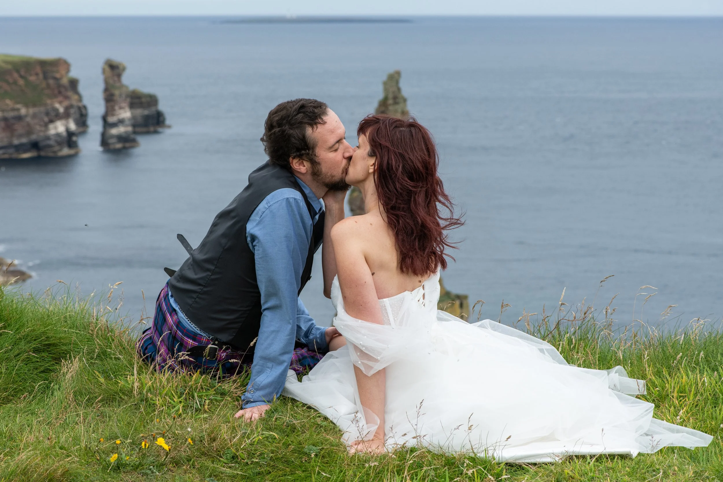 A couple sharing a kiss outdoors on grassy terrain with the sea and rock formations in the background.