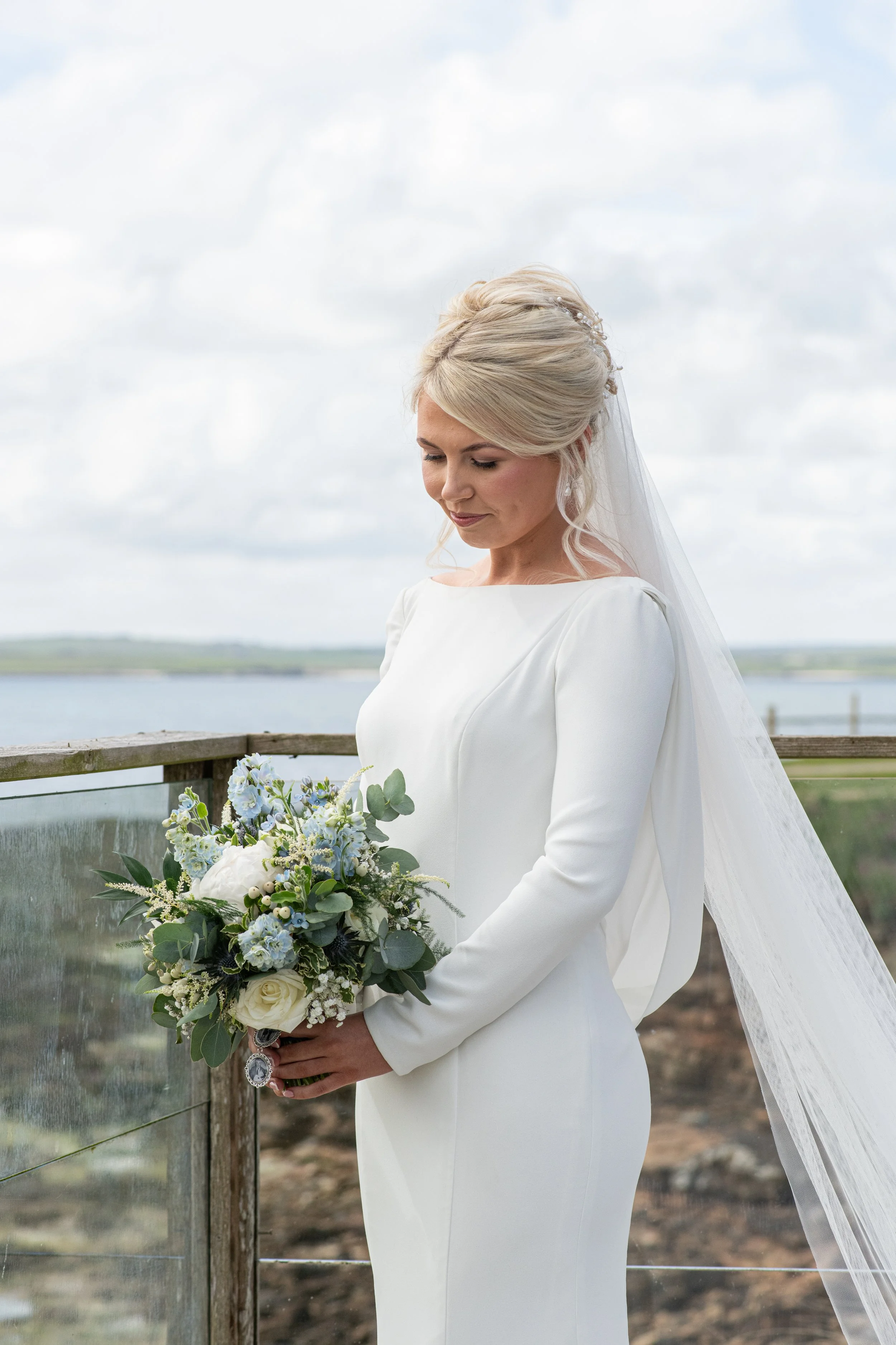 A bride in a white wedding dress with a long veil holds a bouquet of white and blue flowers, standing outdoors near a railing, with a cloudy sky and water in the background.