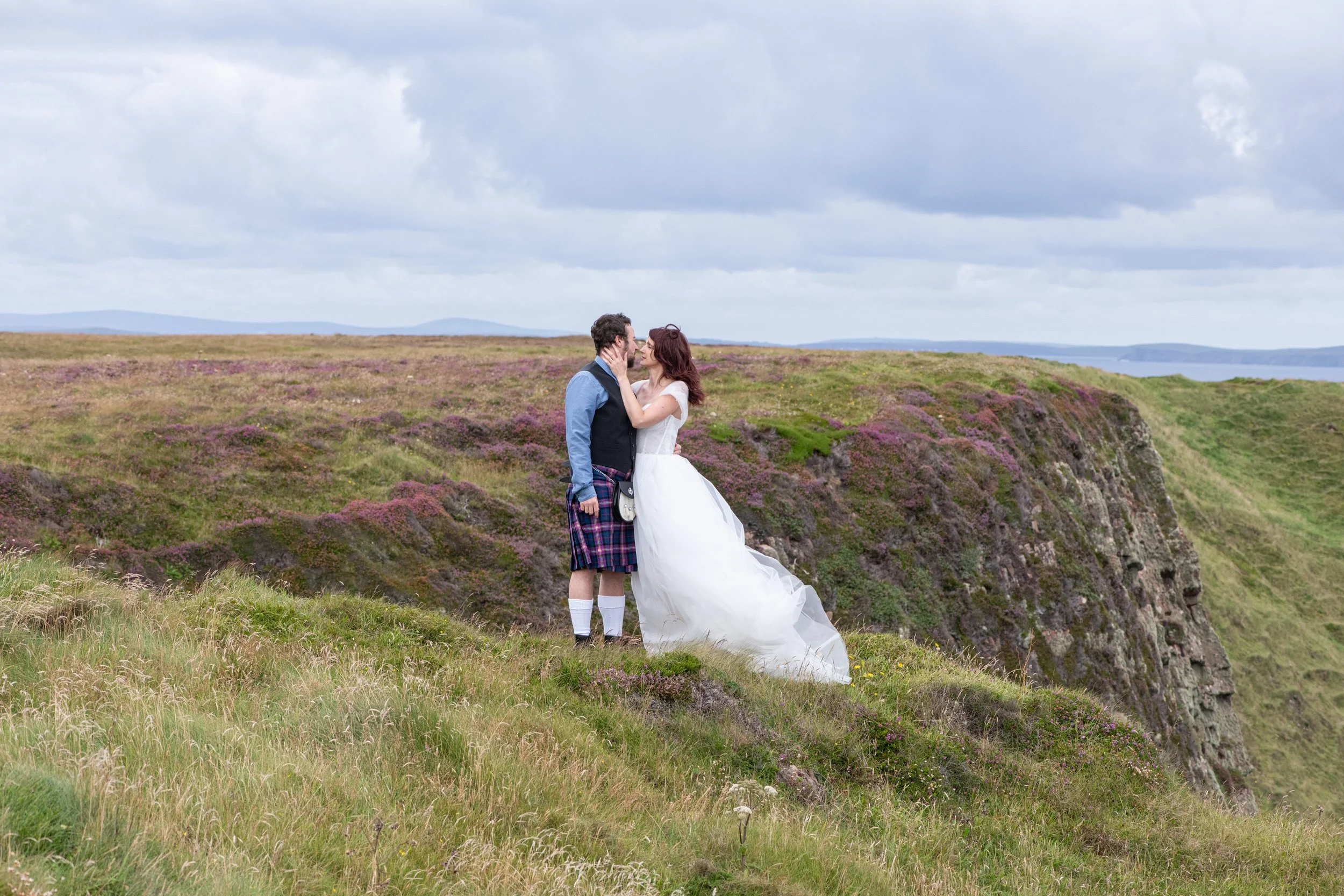 A couple dressed in wedding attire standing on a grassy hillside, about to kiss, with purple wildflowers and cliffs in the background under a cloudy sky.