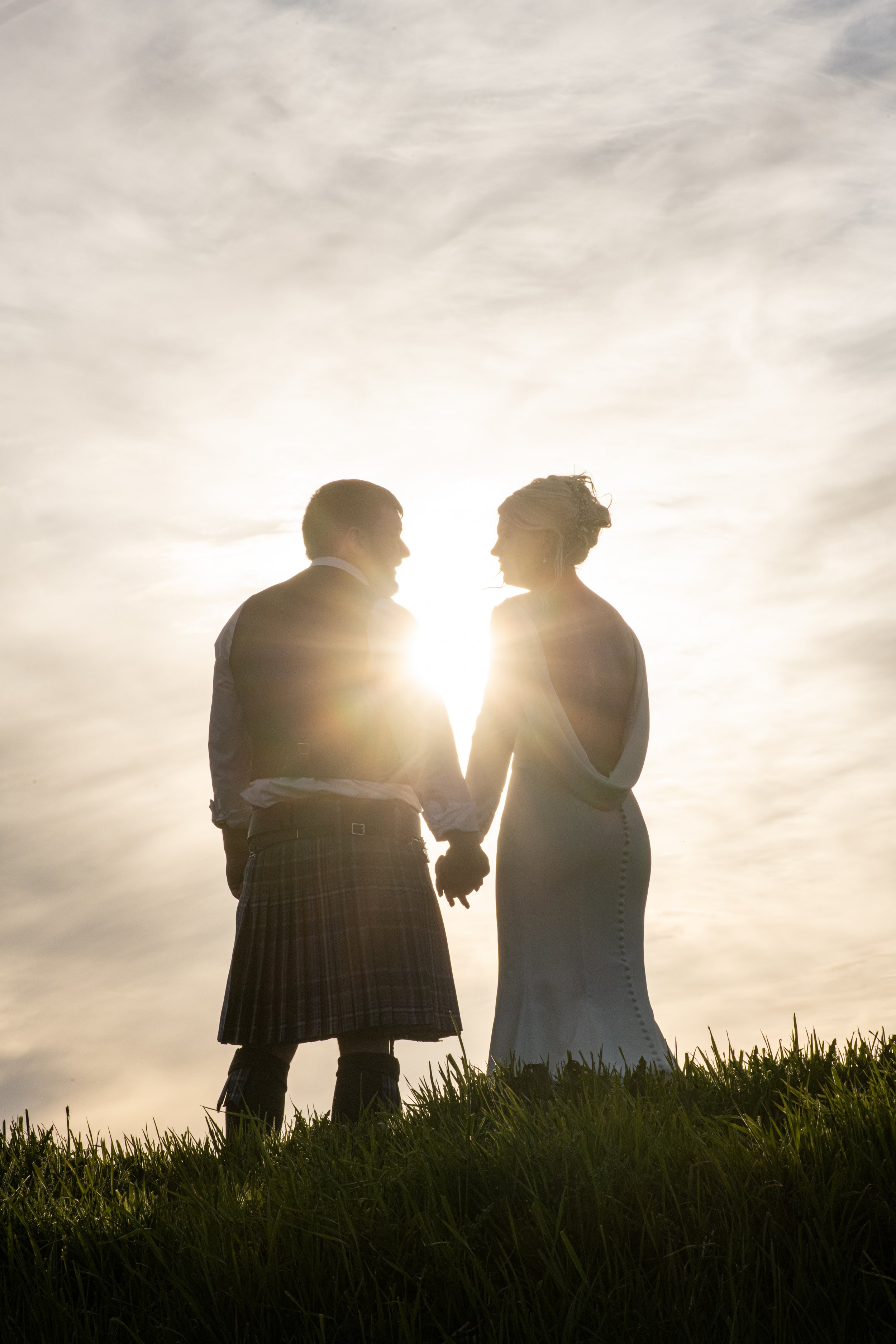 A couple holding hands standing on grass with the sun setting behind them, creating a silhouette effect.
