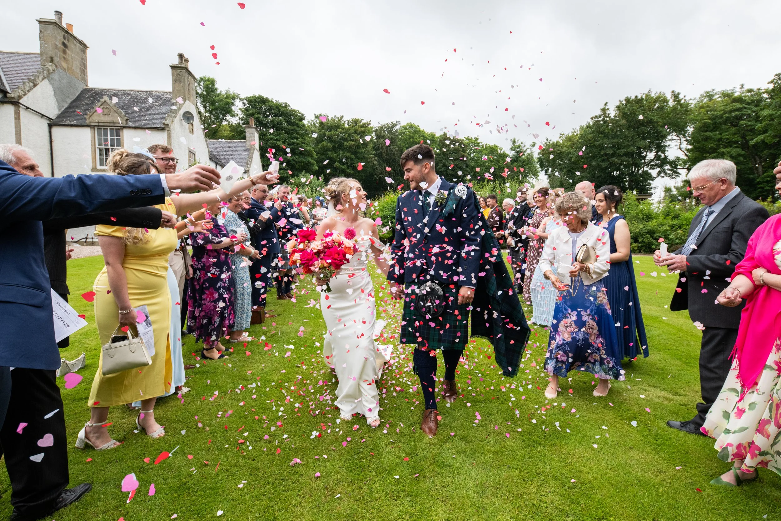 A wedding celebration with a bride and groom walking through a shower of pink and white heart-shaped confetti on a grassy lawn, with guests throwing confetti and smiling.