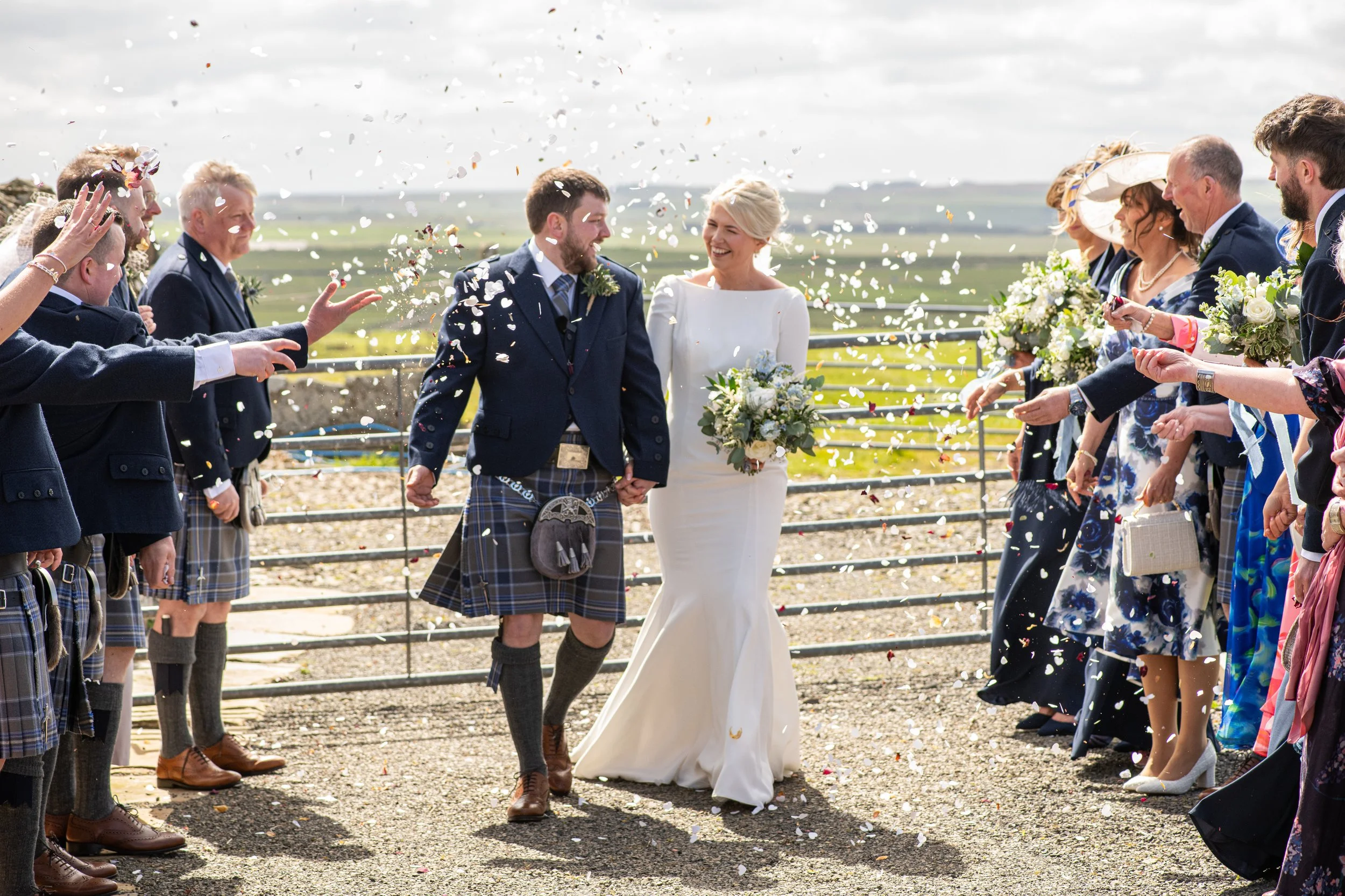 A bride and groom walking hand in hand through confetti, smiling, surrounded by friends and family at an outdoor wedding celebration with a scenic countryside background.
