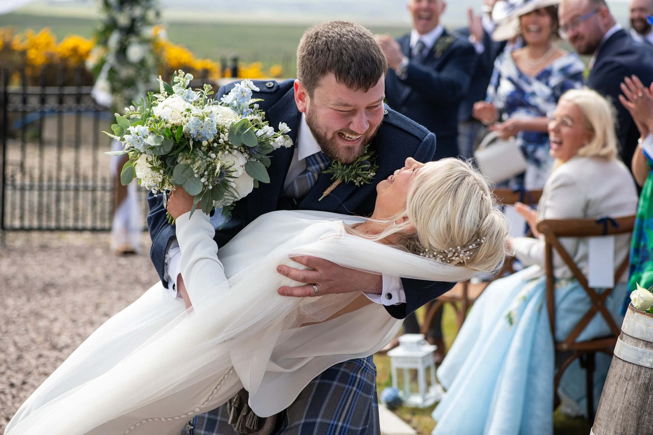 A groom smiling and holding a bride as they dance at an outdoor wedding ceremony surrounded by guests, with a scenic vineyard in the background.