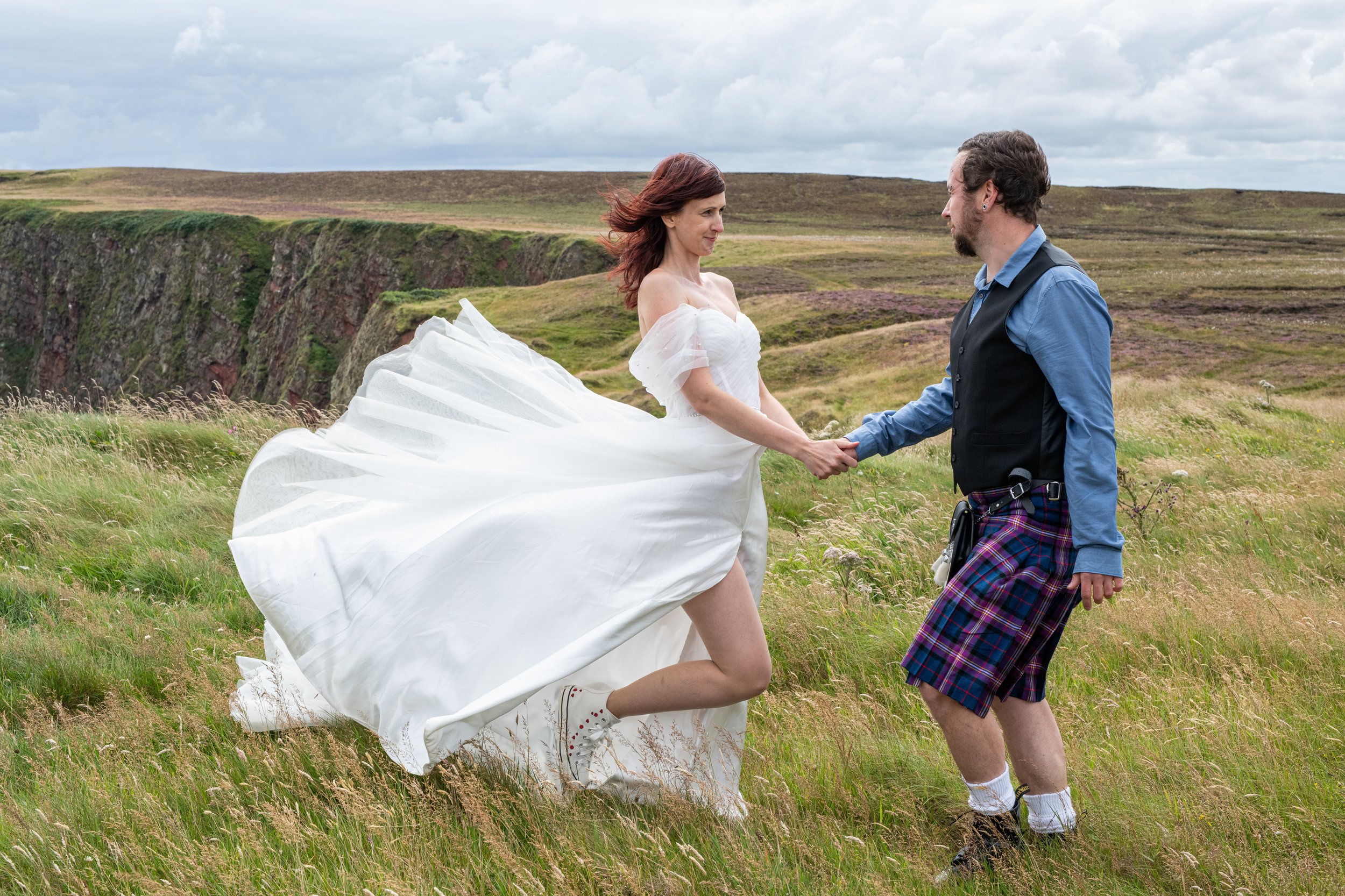 A woman in a wedding dress holding hands with a man in a kilt, standing in a grassy field with cliffs and cloudy sky in the background.