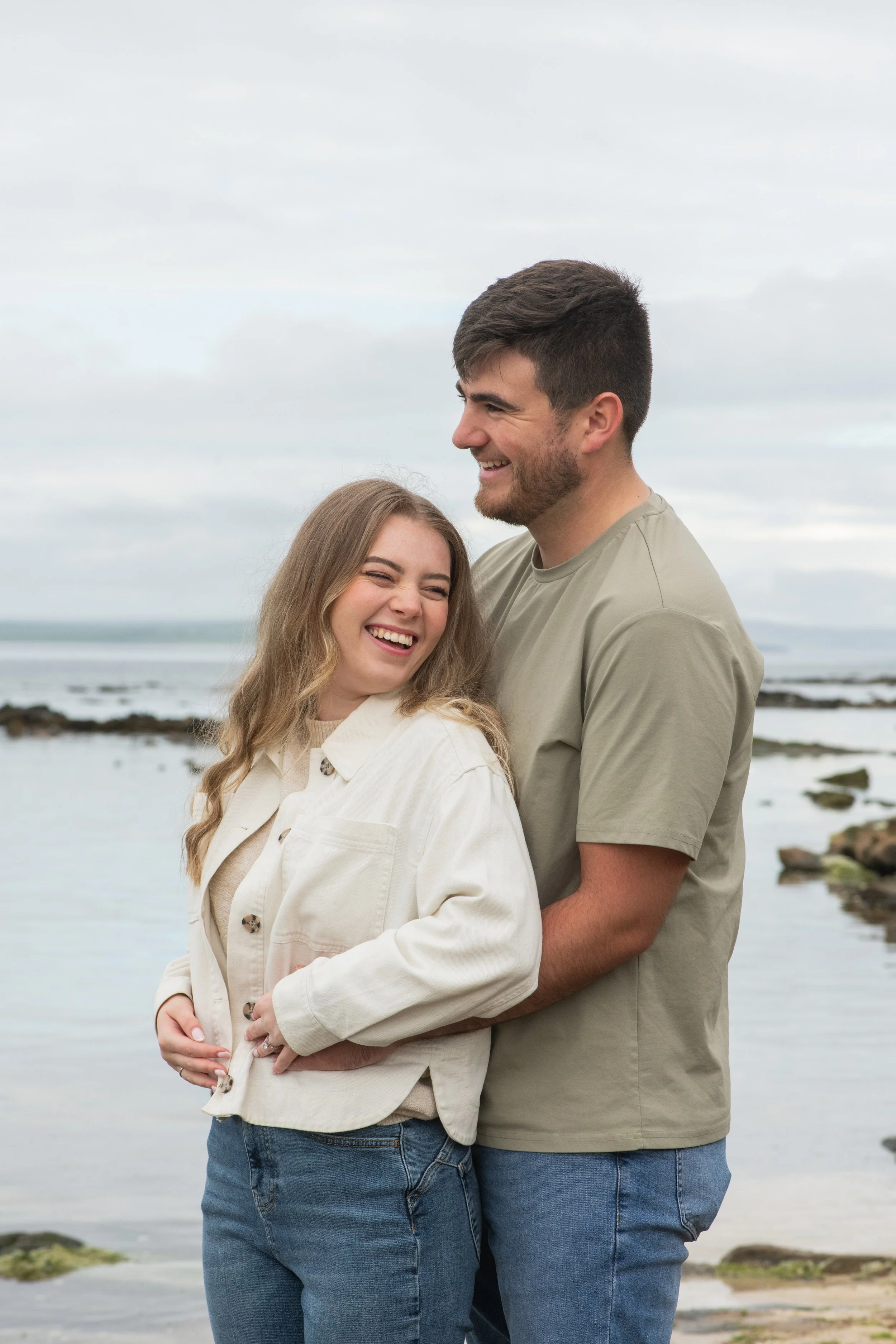 A smiling young couple standing close together outdoors at the beach, with water and rocks visible in the background.