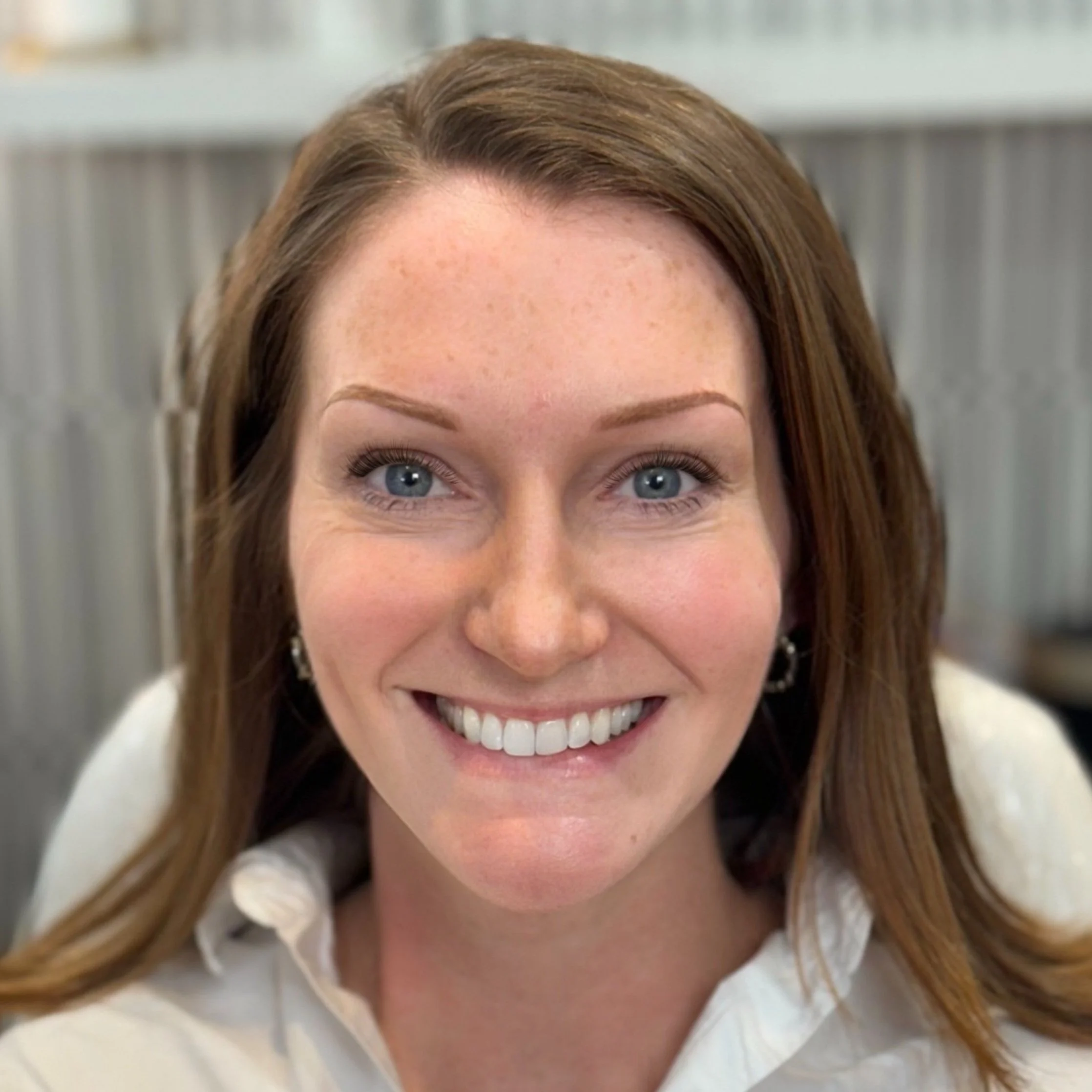 Close-up of a woman with red hair, blue eyes, and a wide smile, wearing a white shirt, in an indoor setting with a blurred background.
