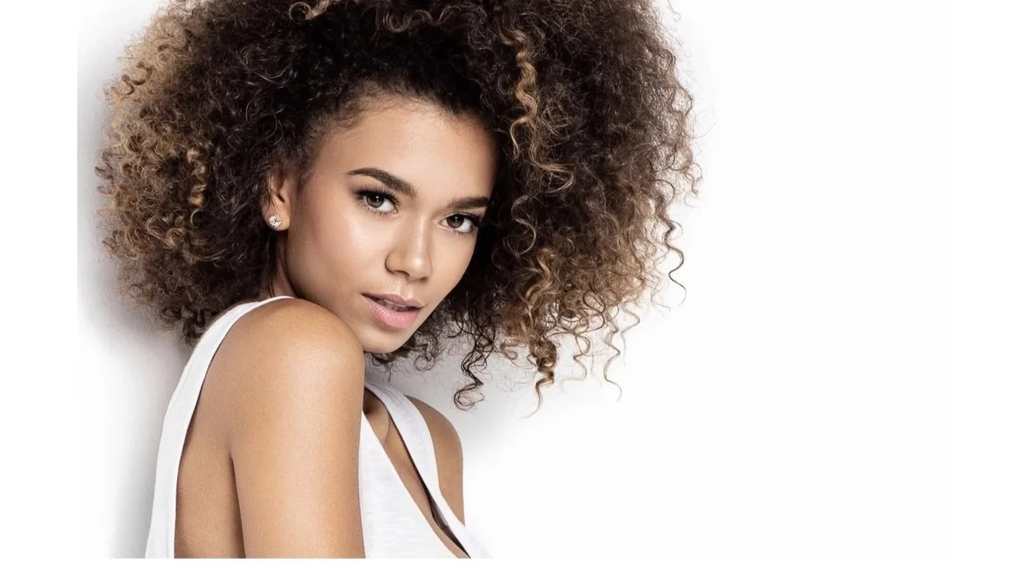 A woman with curly brown hair wearing a white top, looking at the camera with a soft expression, against a white background.