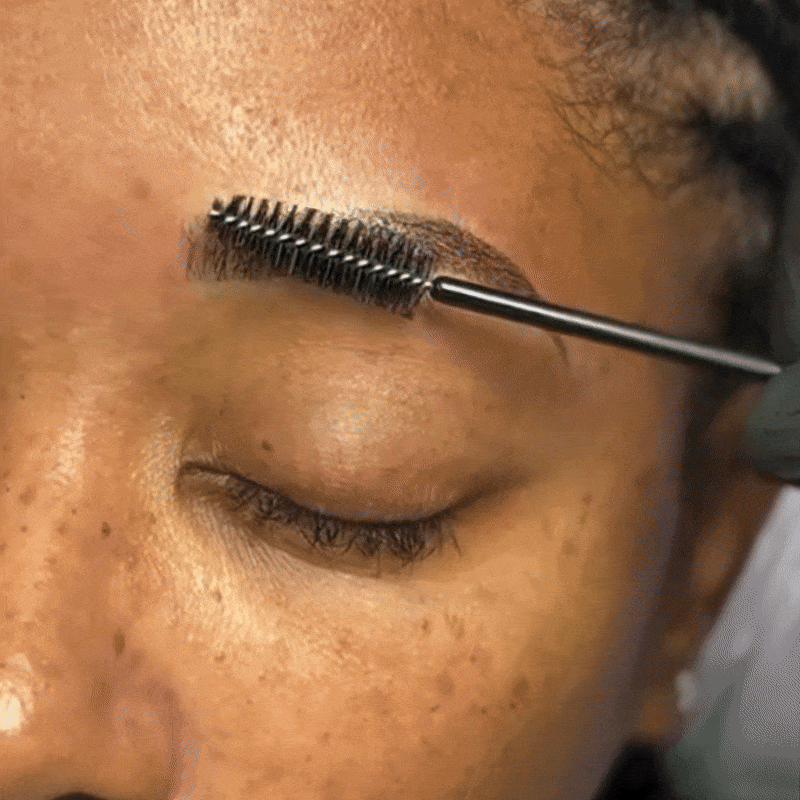 Close-up of a person with their eye closed, applying eyebrow gel with a spoolie brush.