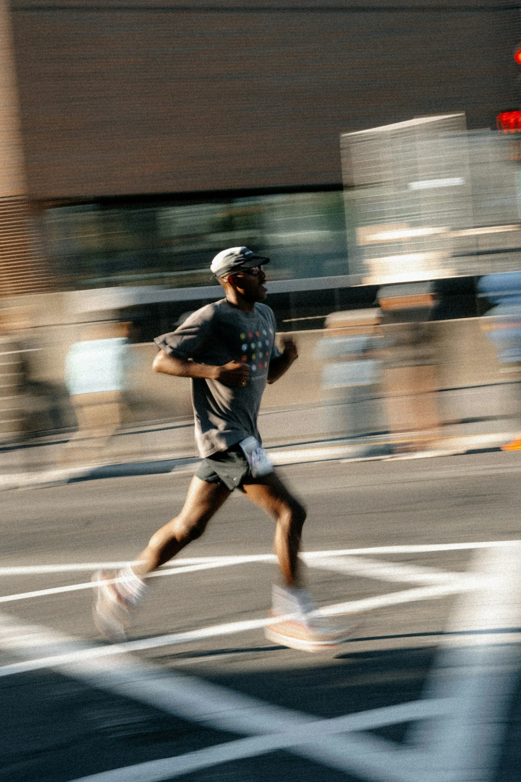 A man running on a city street at sunset, wearing a gray cap, glasses, a dark T-shirt, shorts, and running shoes.