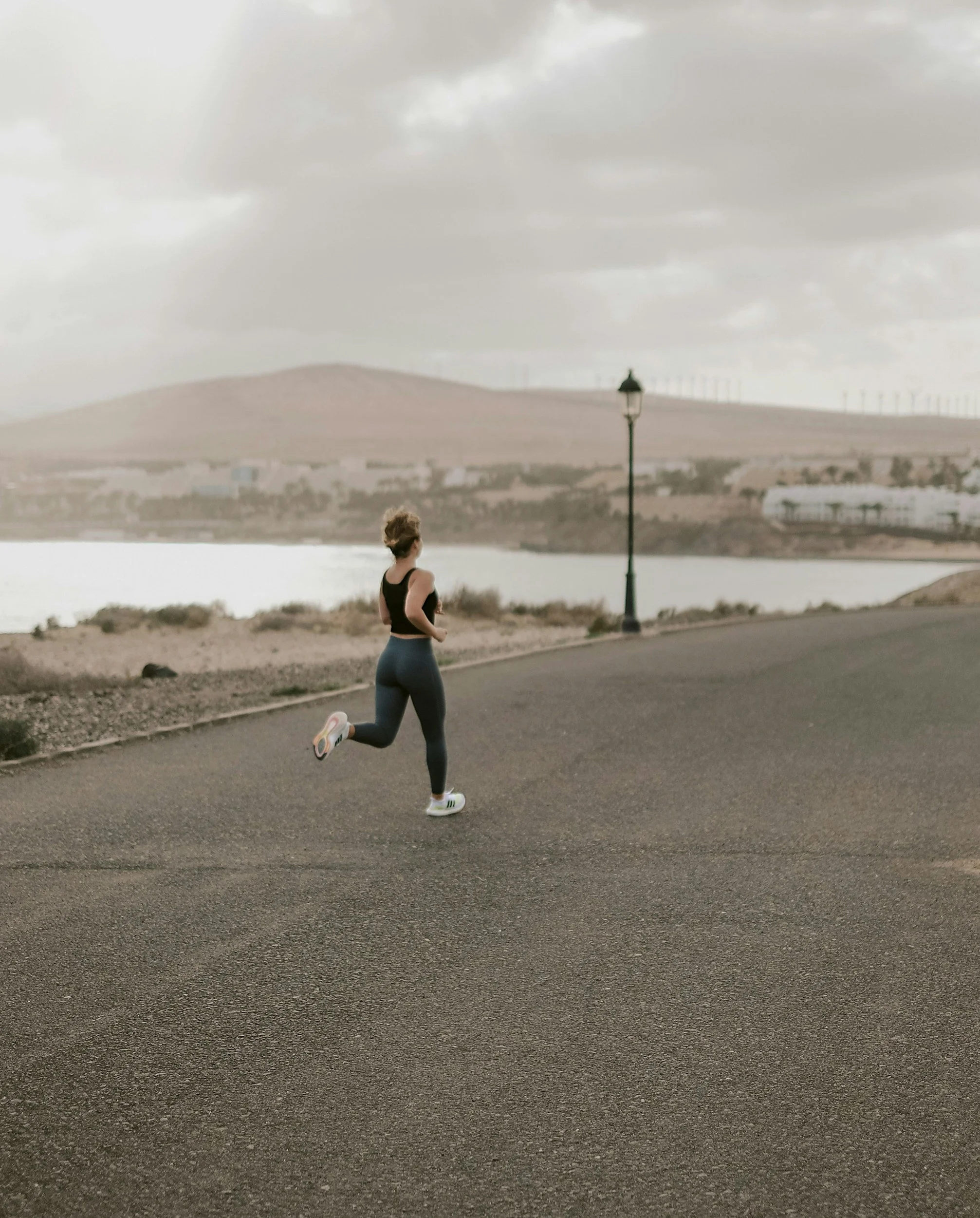 A woman jogging outdoors on a paved road near a calm body of water with hills and wind turbines in the background on a cloudy day.