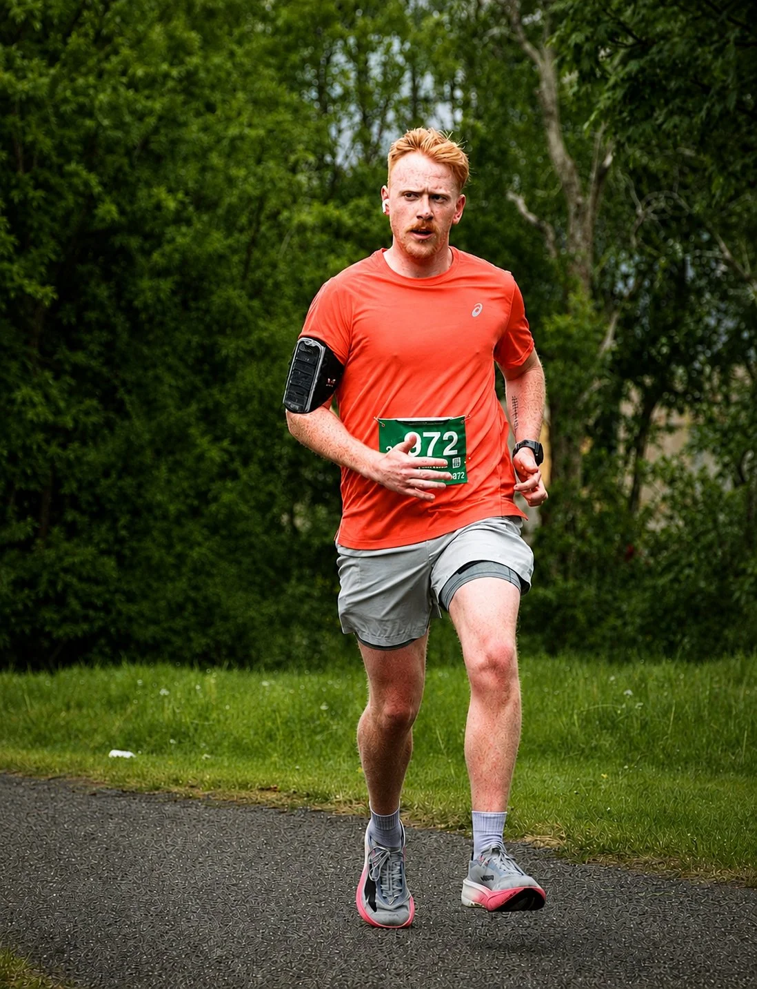 Man running outdoors during a race, wearing a red shirt, beige shorts, gray running shoes, and a watch, with a green bib number 372 on his chest, surrounded by green trees and grass.