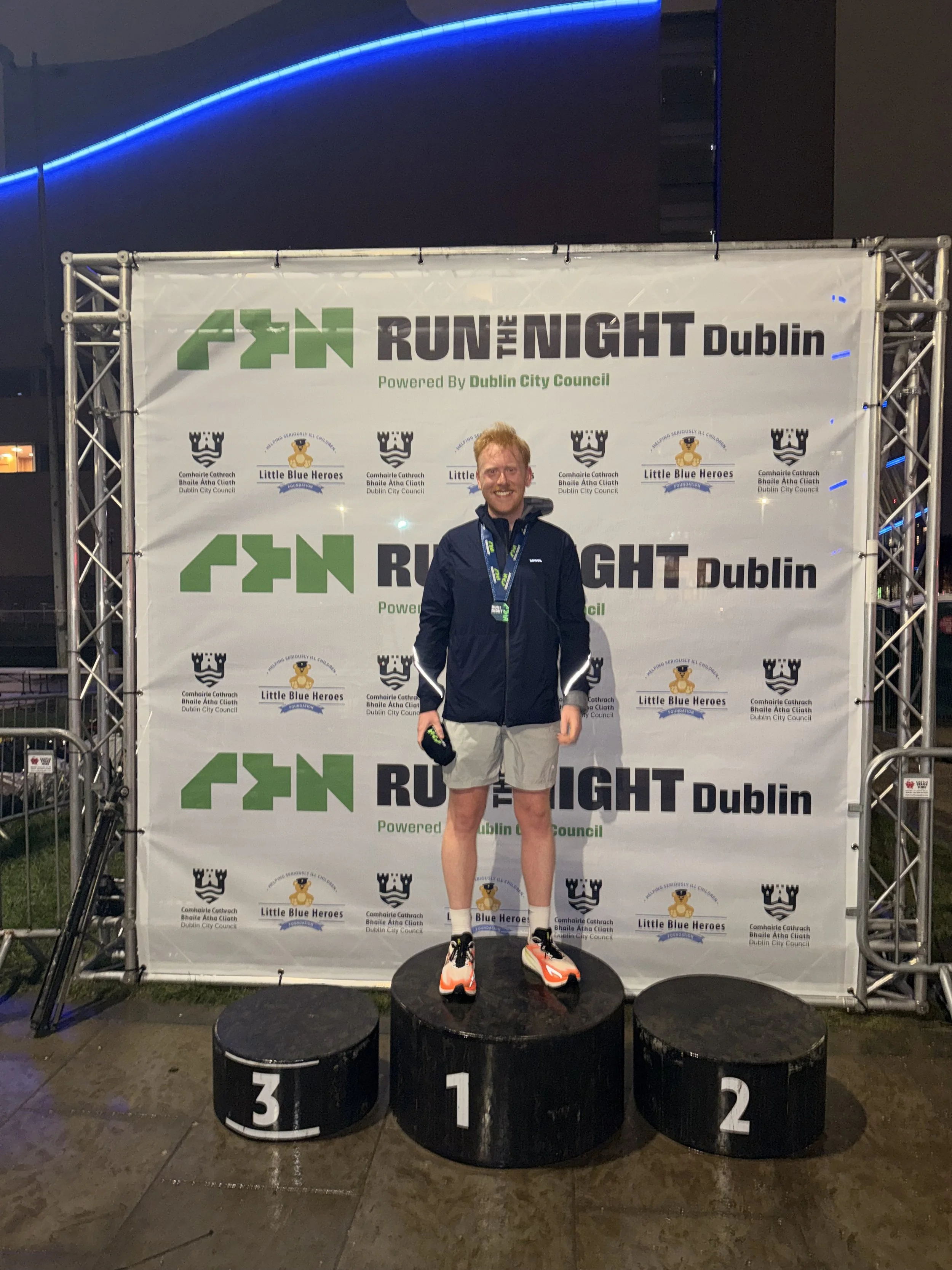 A smiling man standing on the first place podium at a nighttime event, in front of a banner that reads 'Run the Night Dublin,' with logos of Dublin City Council and Little Blue Heroes.