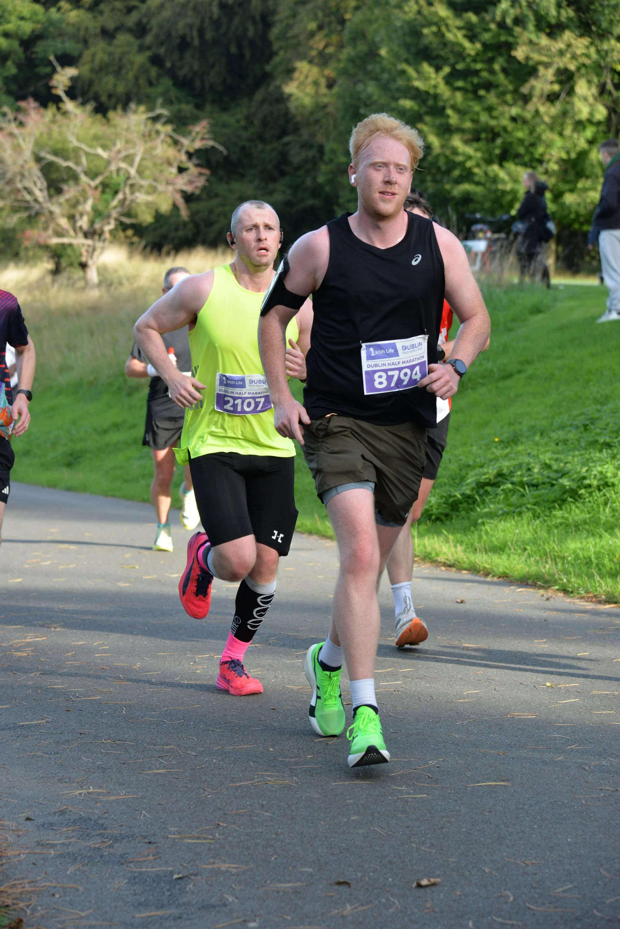 Runners participating in a marathon on a paved path in a park, wearing race bibs and sportswear, with trees and grass in the background.
